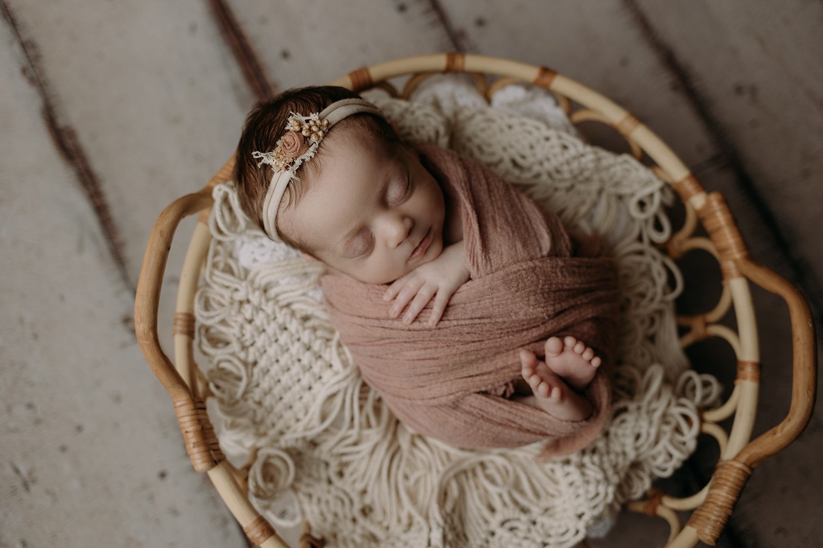 A sleeping baby wrapped in a pink blanket lying in a round wicker basket with a cream-colored knit blanket inside, wearing a floral headband.