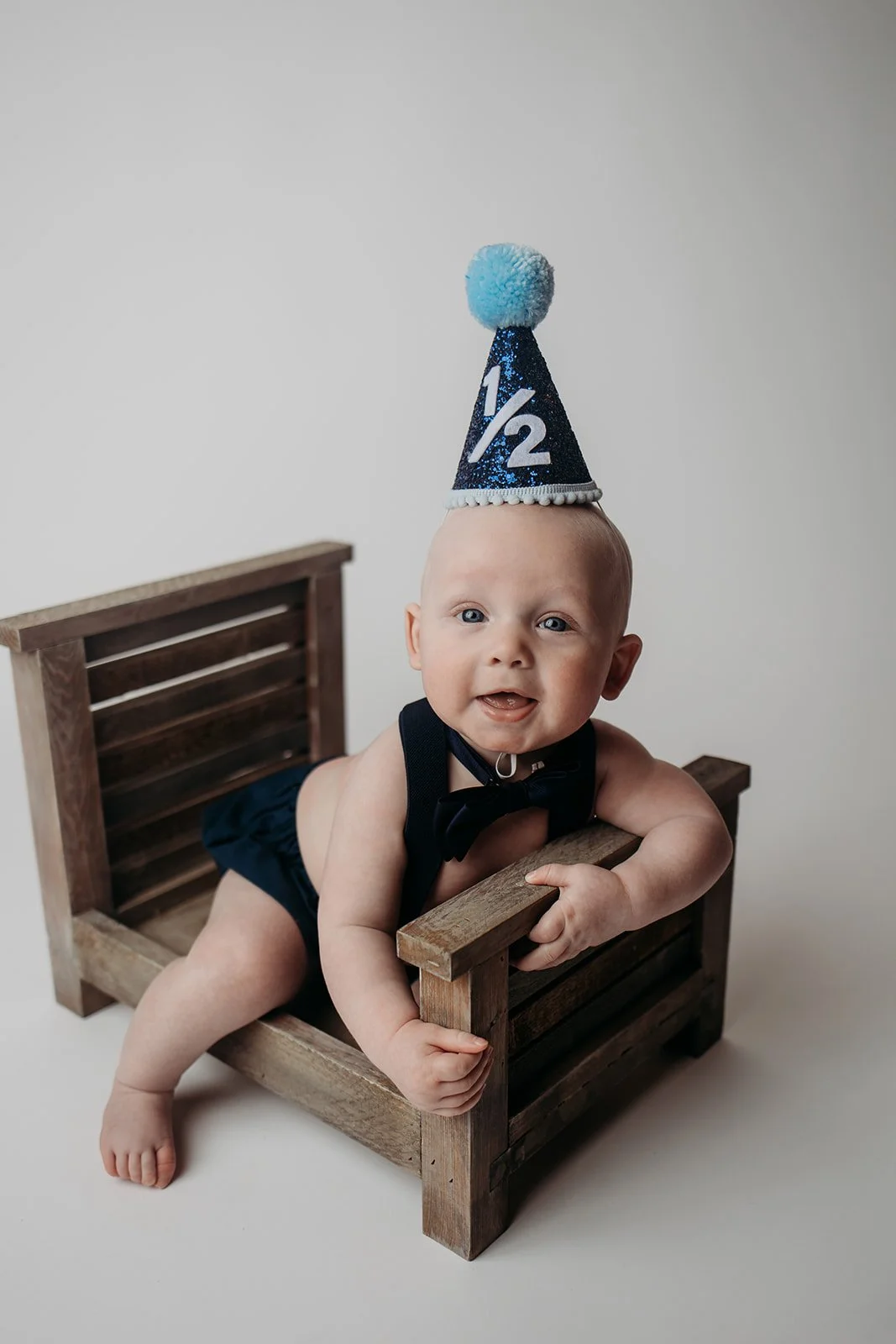 Baby wearing a birthday hat with '1/2' on it, lying on a small wooden bed, holding the edge, and smiling.