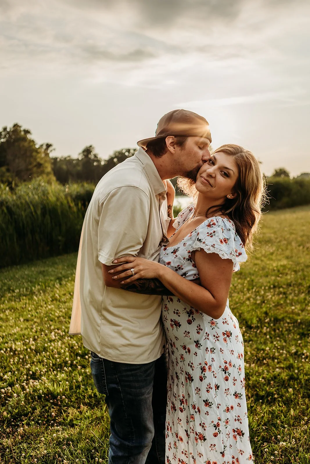 A couple standing in a field during sunset, with the man kissing the woman on the temple and the woman smiling at the camera, the woman wearing a white floral dress and the man wearing a beige shirt and cap.