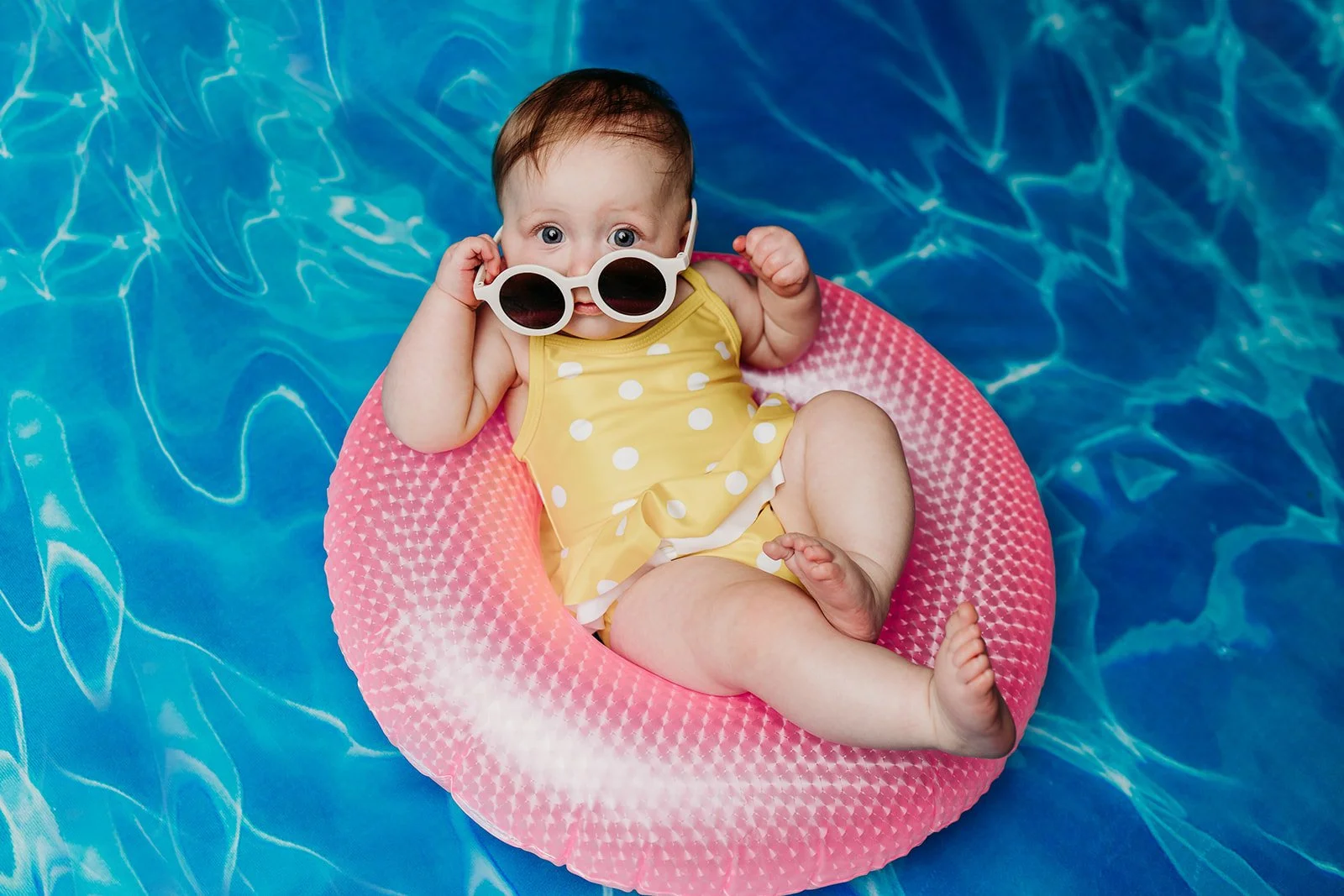 A baby wearing sunglasses and a yellow polka dot swimsuit, sitting on a pink pool float in a blue swimming pool.