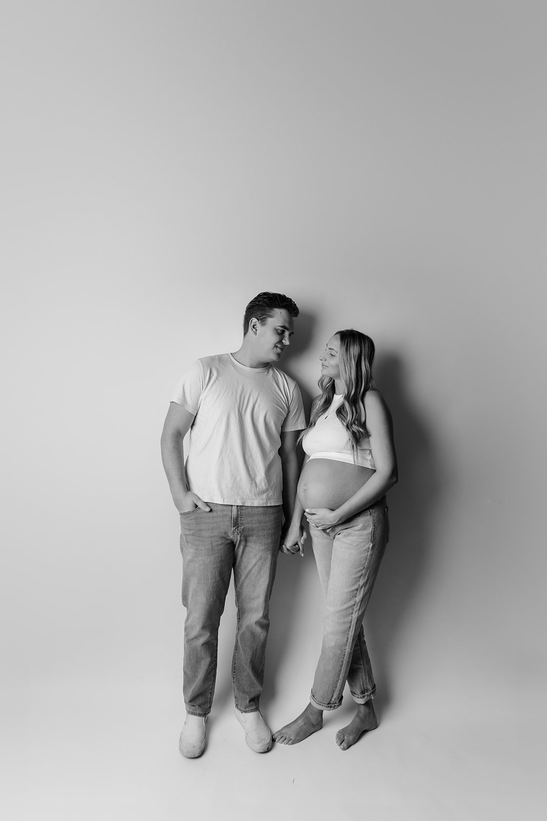 A smiling pregnant woman and a man holding hands and looking at each other in a studio with a plain background.