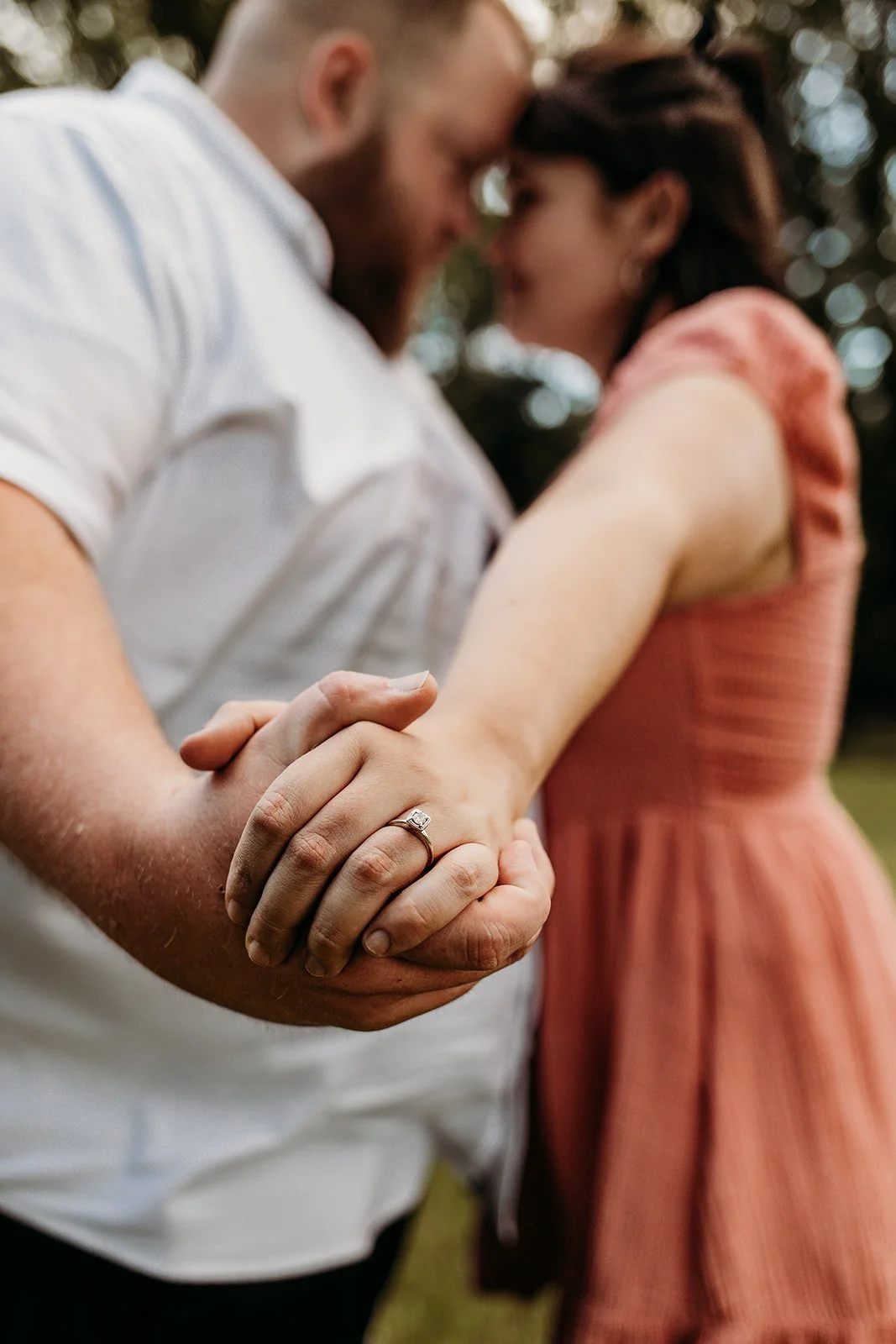A close-up of a couple holding hands, with a focus on the woman's wedding ring, while they lean their foreheads together in a blurred outdoor background.