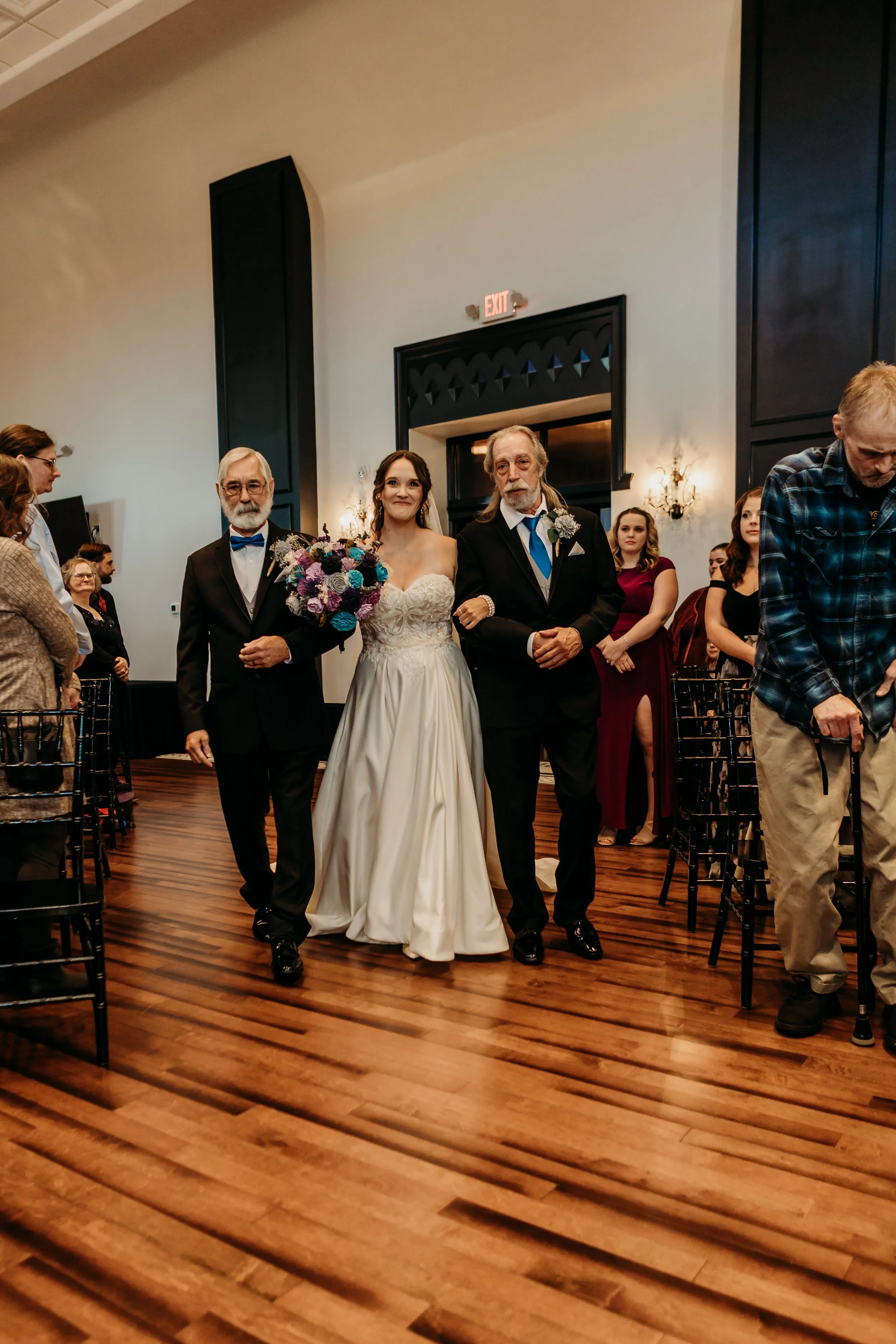 A bride walking down the aisle with her parents in a wedding ceremony inside a decorated hall, with guests seated on either side.