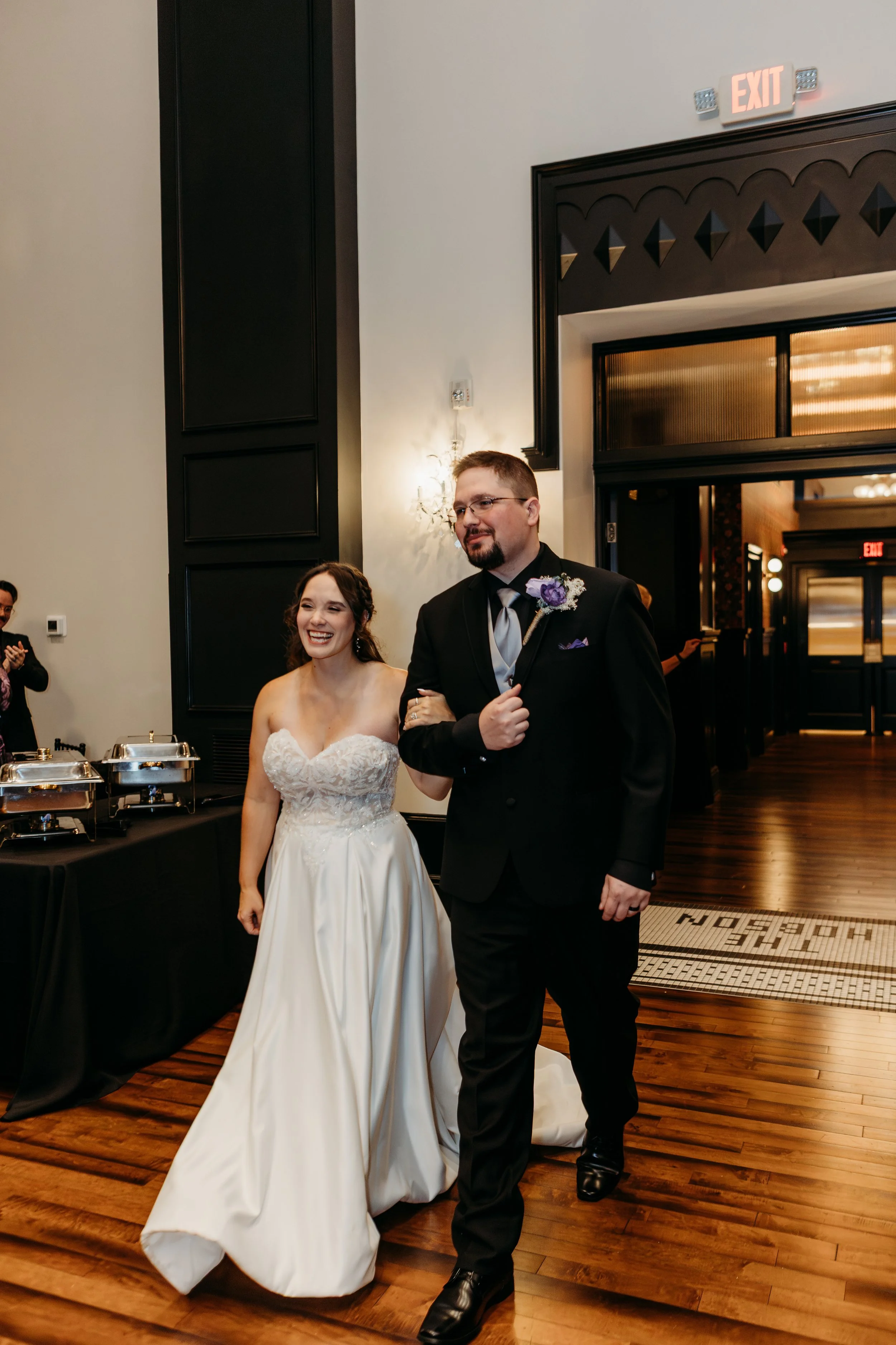 A bride and a groom walking arm-in-arm at their wedding reception. The bride is wearing a white strapless wedding dress, and the groom is dressed in a black tuxedo with a purple boutonniere. They are smiling and appear happy. There are guests in the 