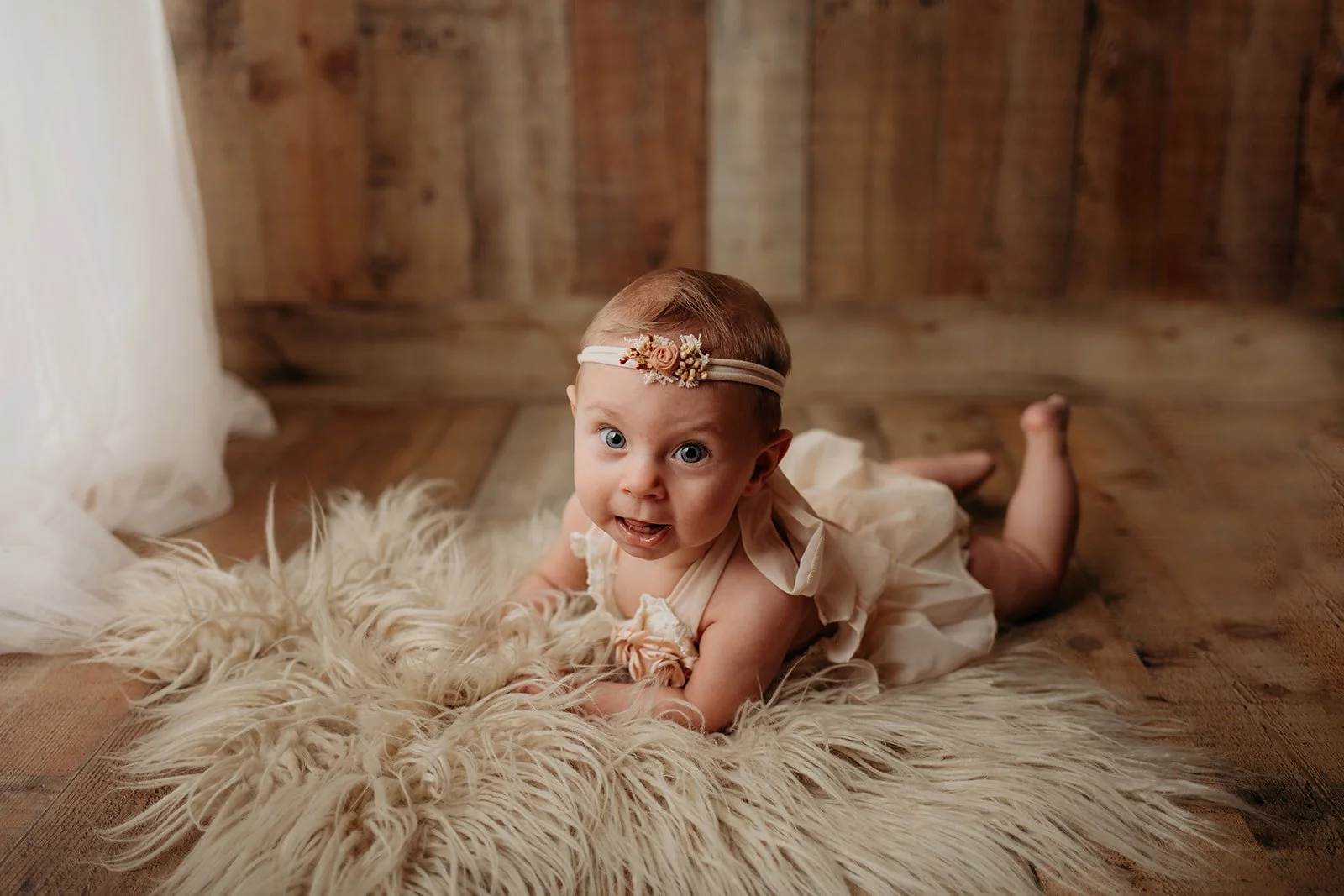 A baby girl lying on a fluffy cream-colored rug, looking at the camera with wide eyes, wearing a cream dress and a floral headband, with a wooden wall in the background.