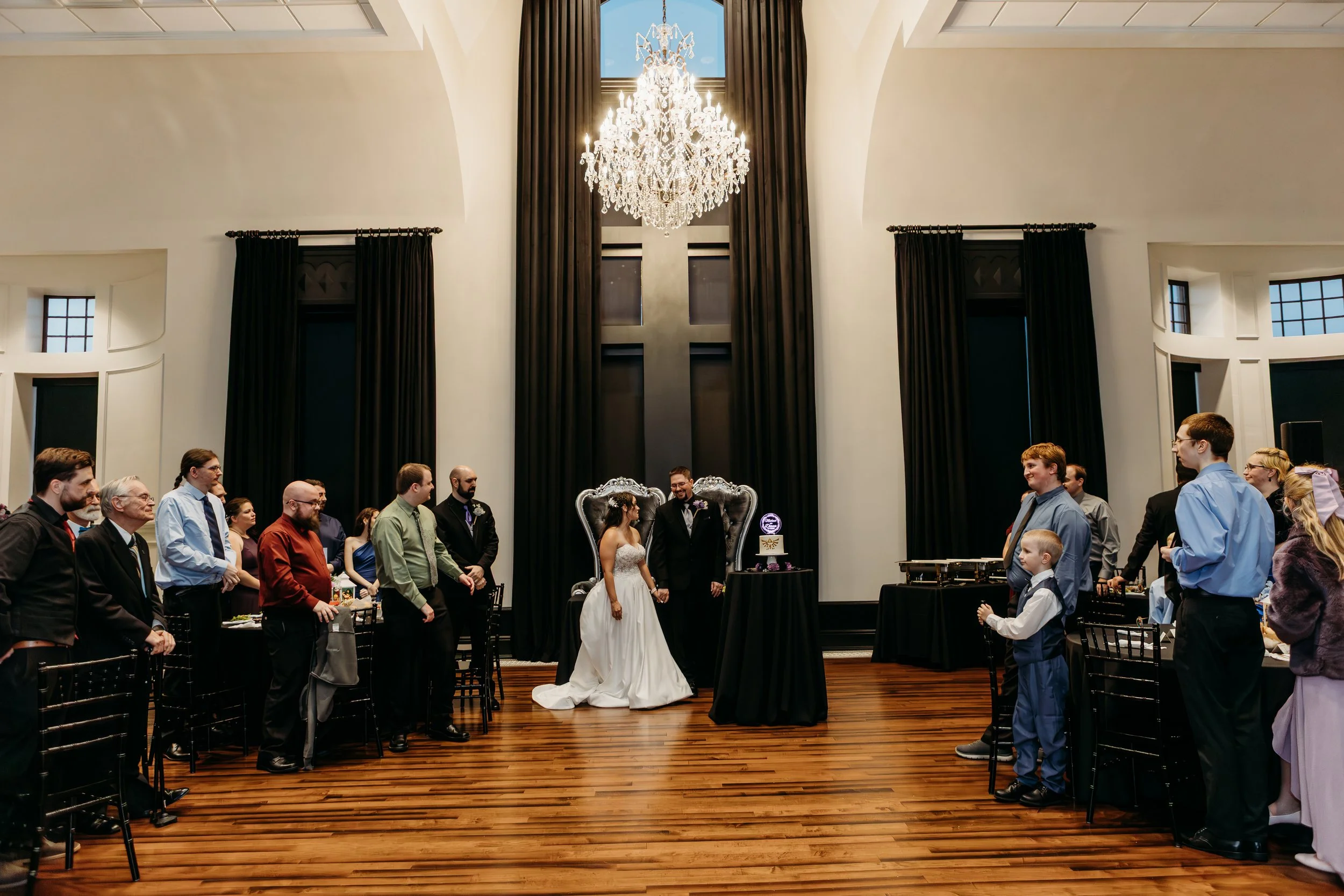 A wedding ceremony taking place in an elegant hall with a high ceiling, chandelier, and dark curtains. The bride and groom stand in front of a large cross with guests gathered on either side.