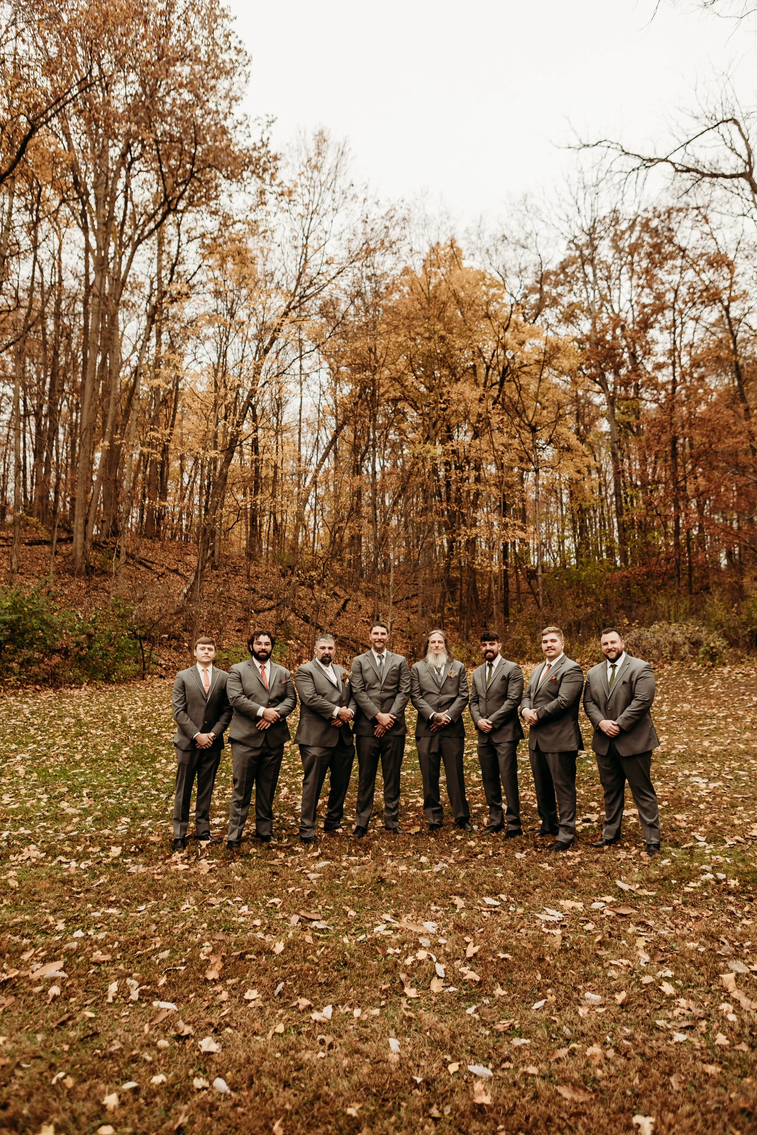 A group of seven men in suits standing outdoors on a fall day with trees and leaves in the background.