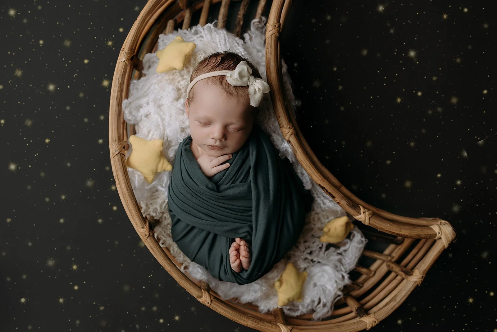 A newborn baby sleeping in a wooden basket, wrapped in dark green cloth, with a cream headband and bow, surrounded by yellow star-shaped plush toys, on a black background with star-like specks.