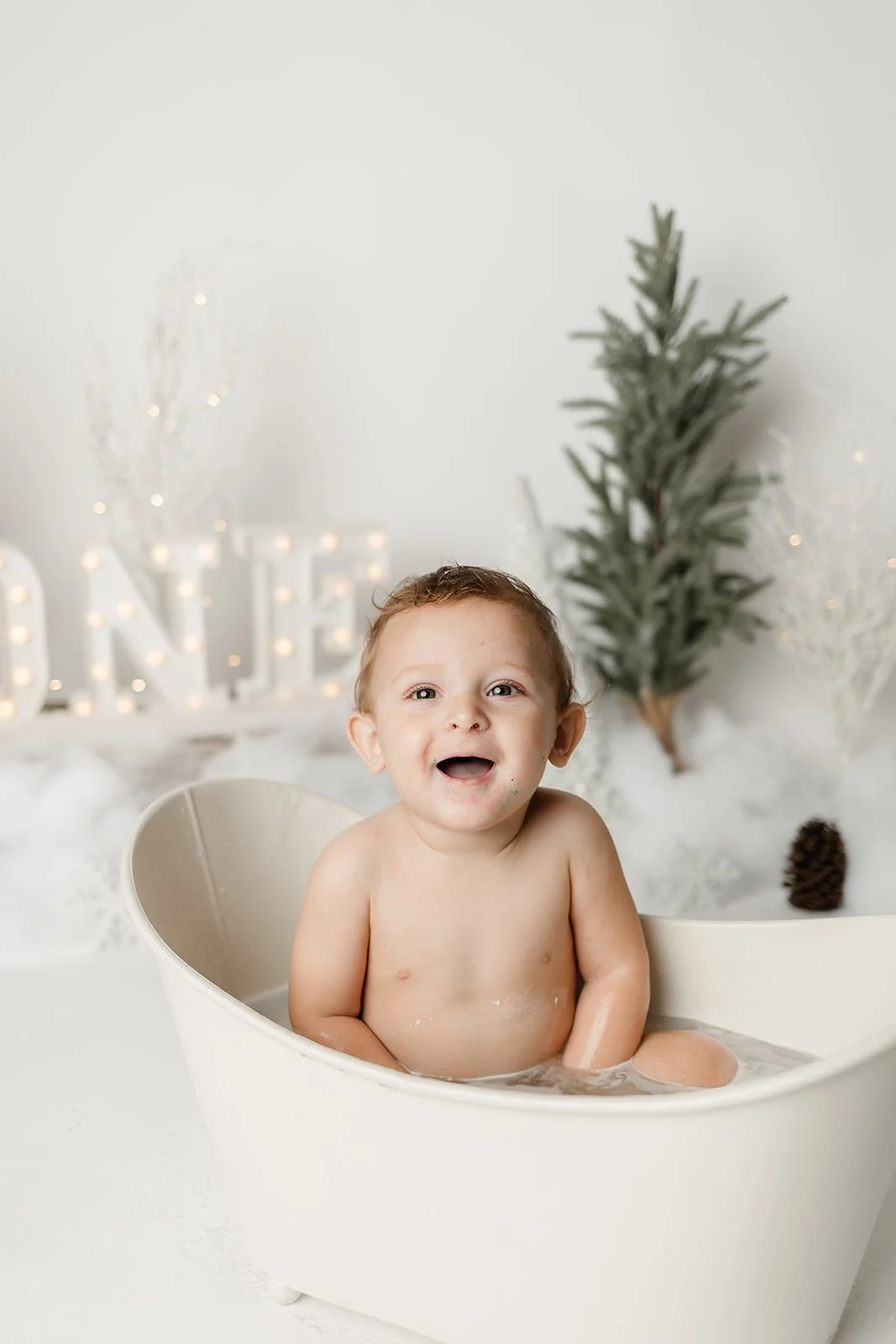 A baby sitting in a white bathtub, smiling and looking at the camera, with a festive holiday background including a small decorated Christmas tree, pinecones, and illuminated decorative letters spelling 'NOEL'.