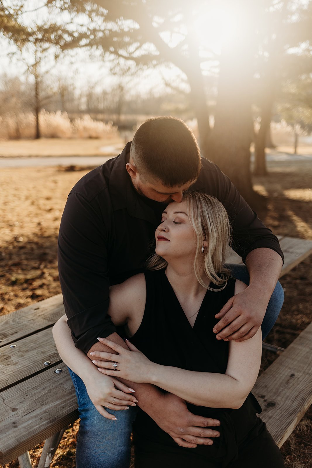 A couple embraces on a park bench during sunset, with a man kissing a woman on the forehead.