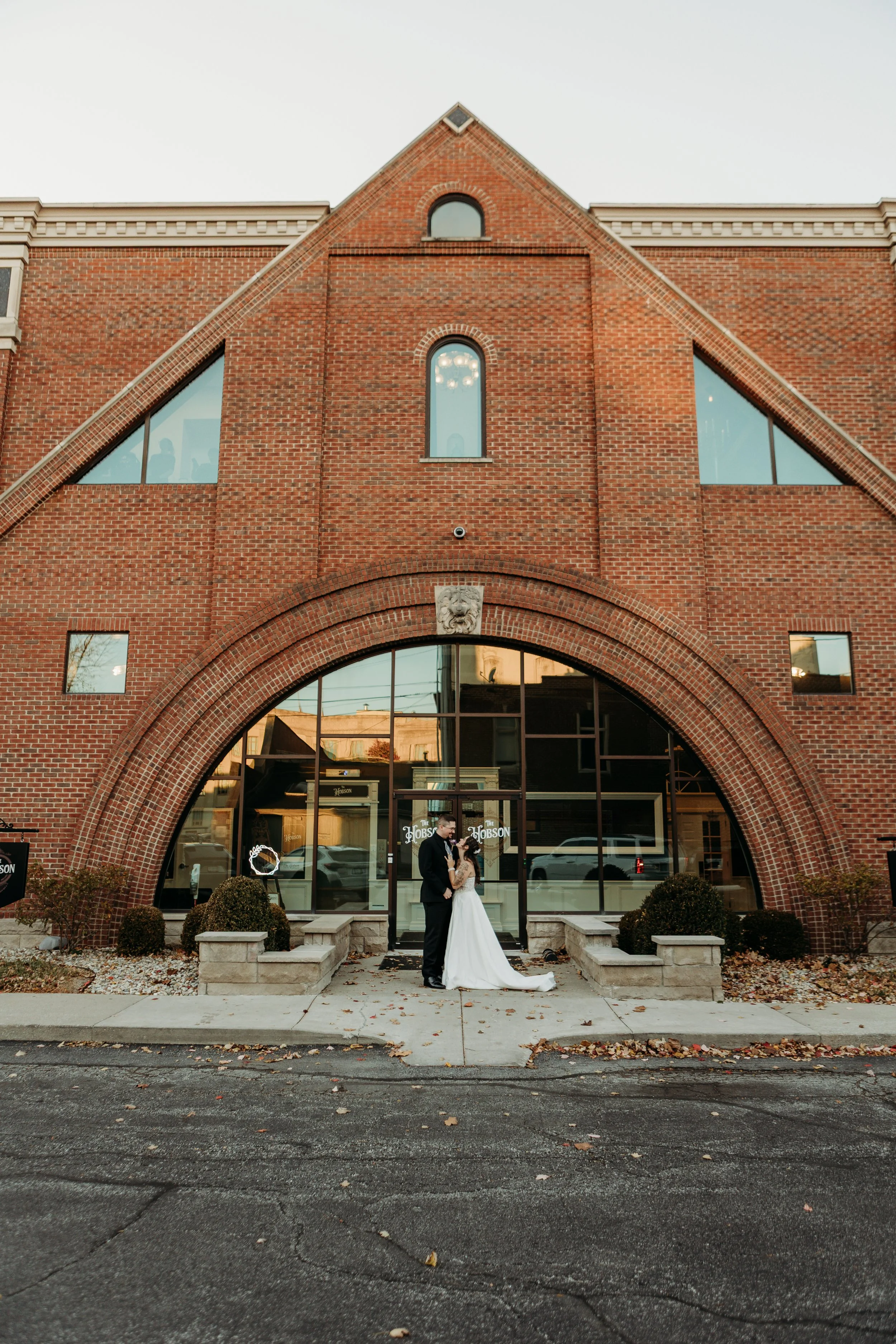 A bride and groom standing close together outside a large brick building with arched windows and a glass entrance, during sunset.