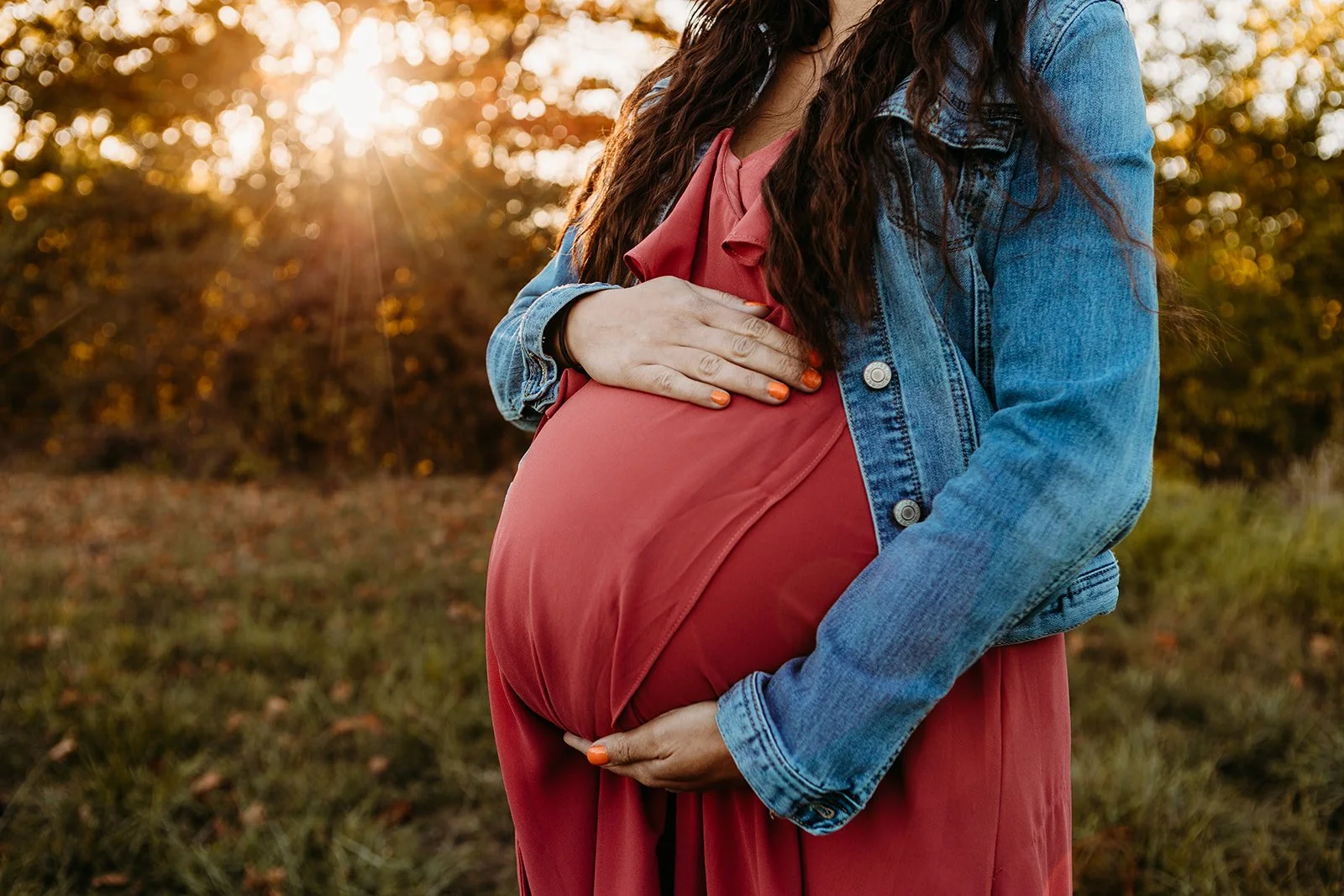 A pregnant woman wearing a red dress and denim jacket is holding her belly outdoors during sunset, with trees in the background.