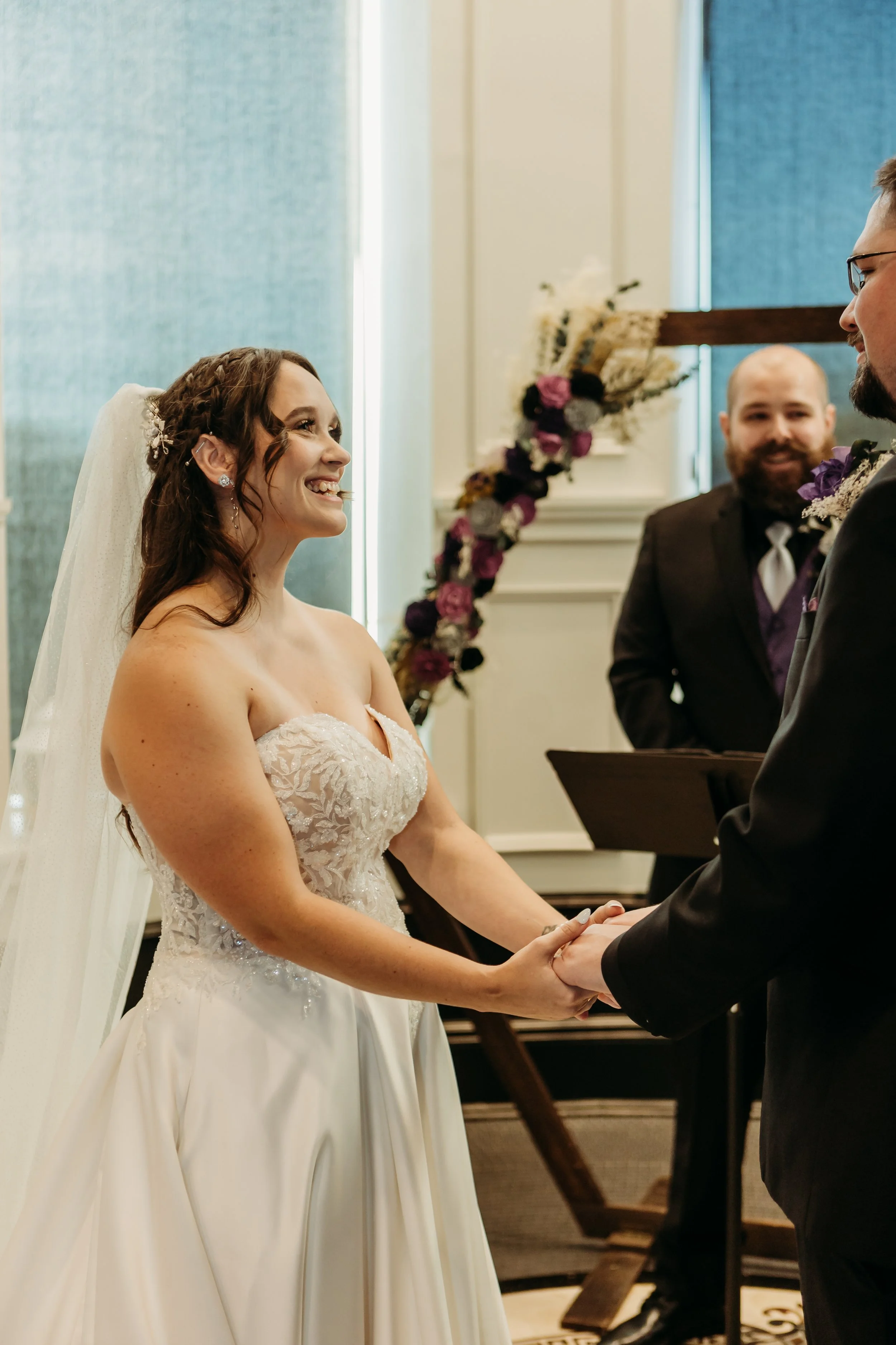 A bride and groom holding hands during their wedding ceremony, smiling at each other, with an officiant standing behind them and floral decorations in the background.