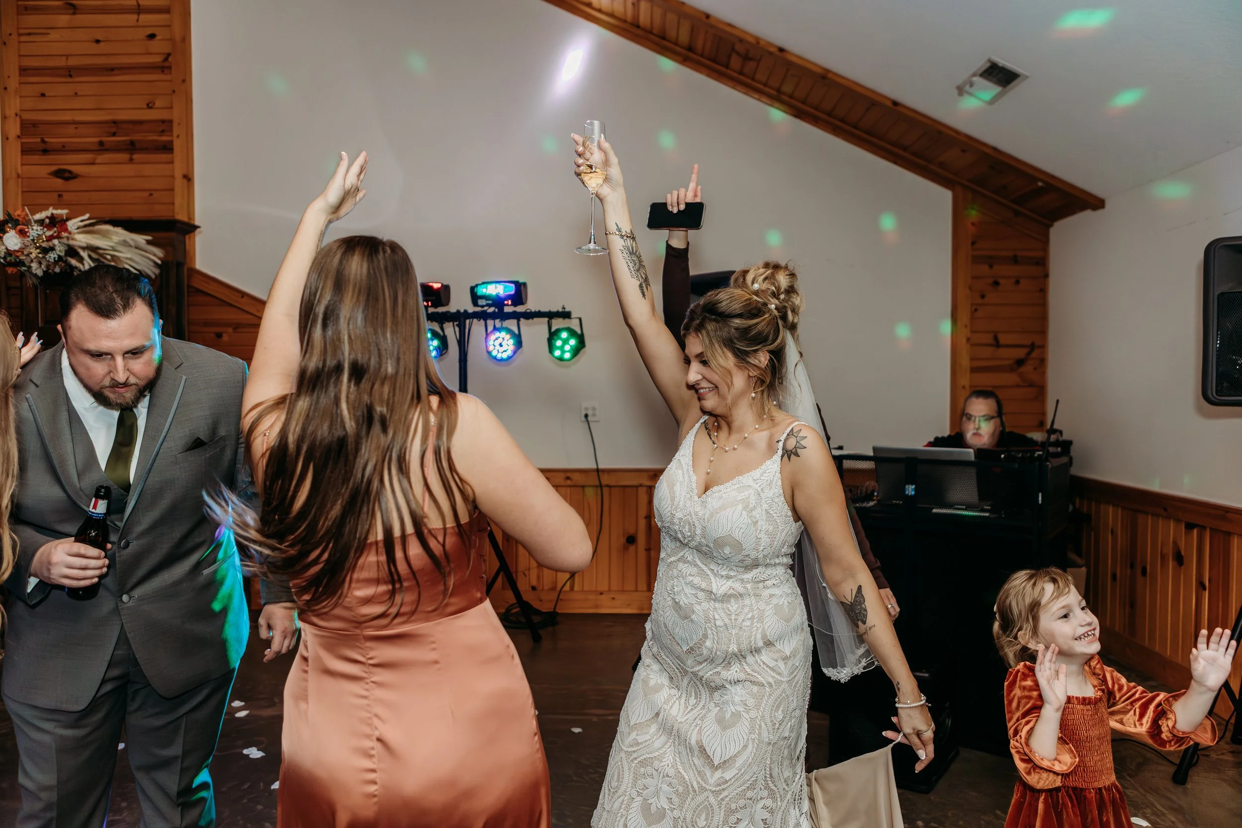 People dancing at a celebration with colorful lights, including a bride in a white wedding gown holding a glass of champagne, and a young girl in a rust-colored dress clapping and smiling.