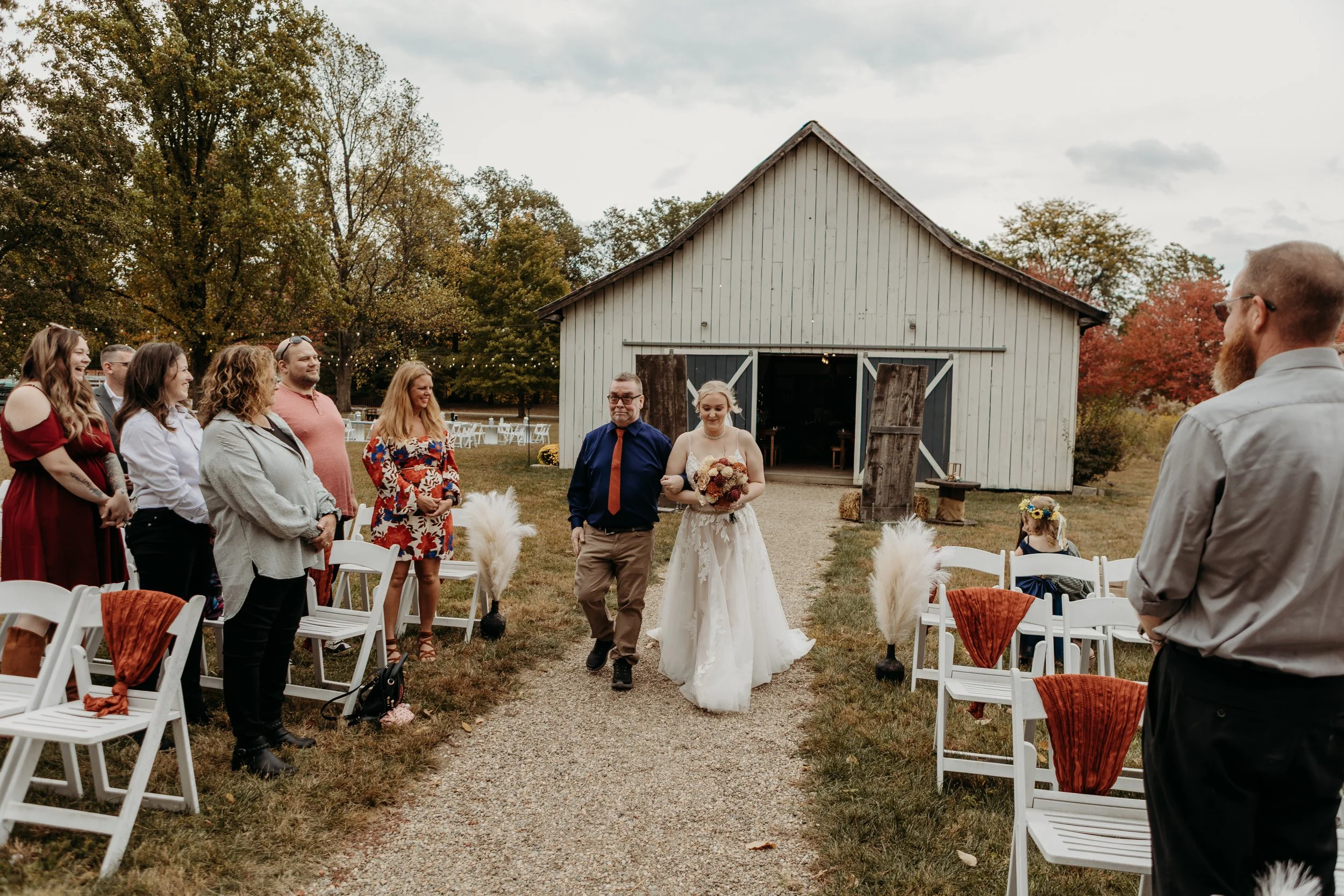 Bride walking down the aisle with a man at a rustic outdoor wedding ceremony, surrounded by seated guests and decorated with white chairs and fall foliage.