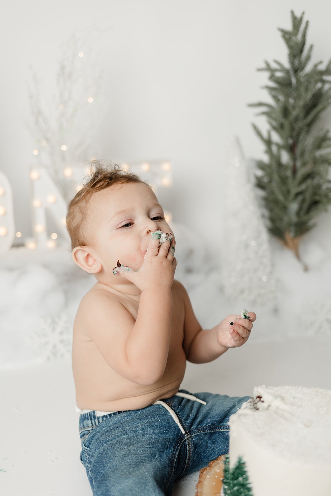 A young boy with no shirt and jeans is eating cake with white frosting, decorated with Christmas-themed sprinkles, against a white background decorated with Christmas trees and lights.