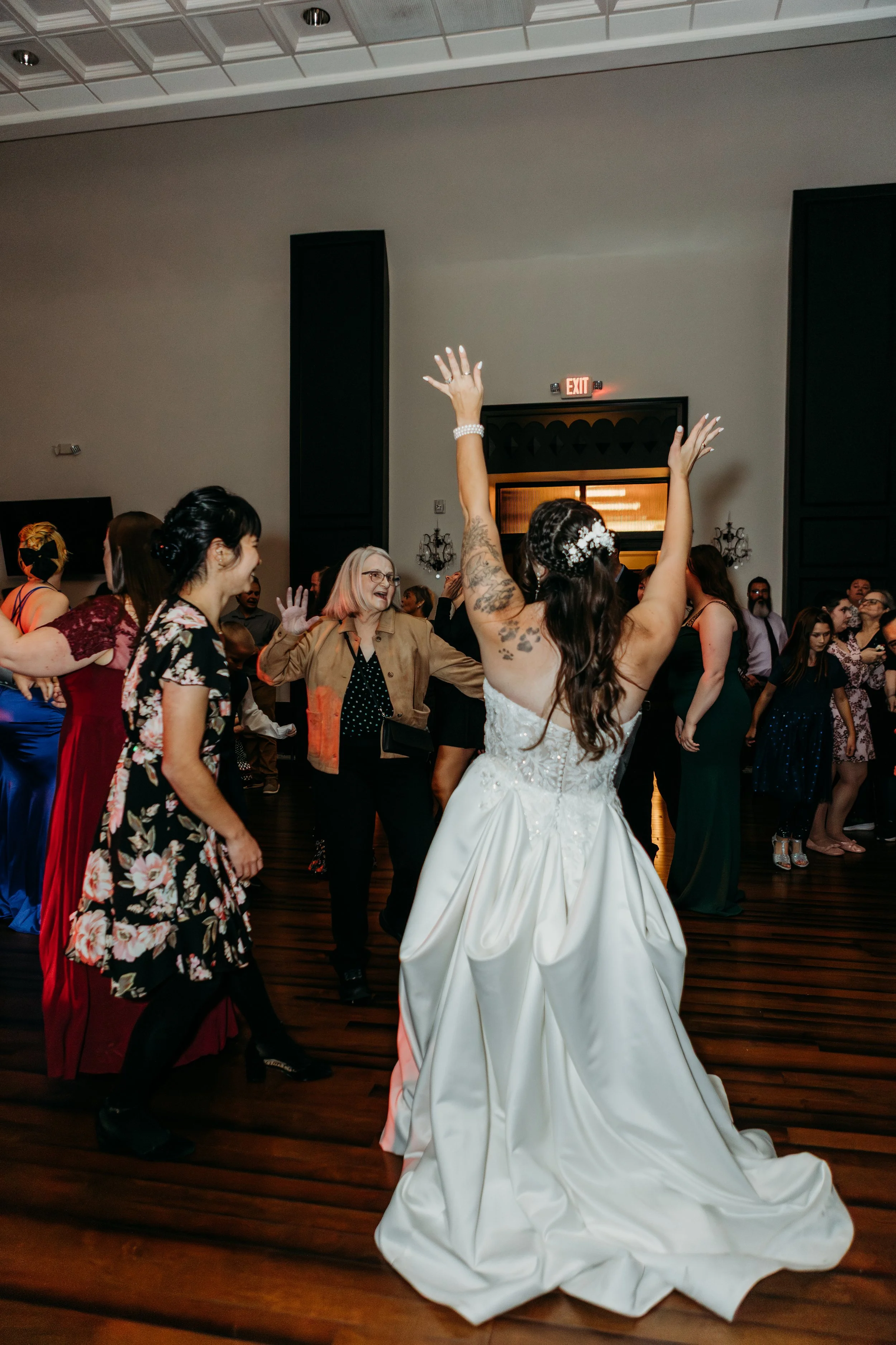 Bride dancing with guests at a wedding reception in a ballroom with wooden floors and gray walls.