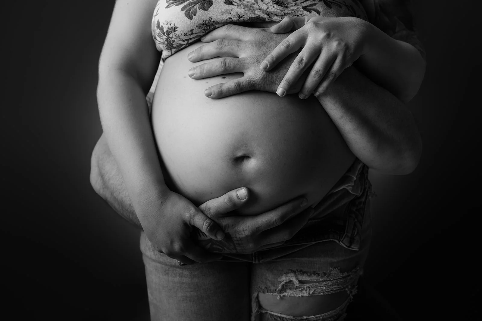 A pregnant woman holding her belly with both hands, with a man's hands also gently resting on her belly, in black and white.