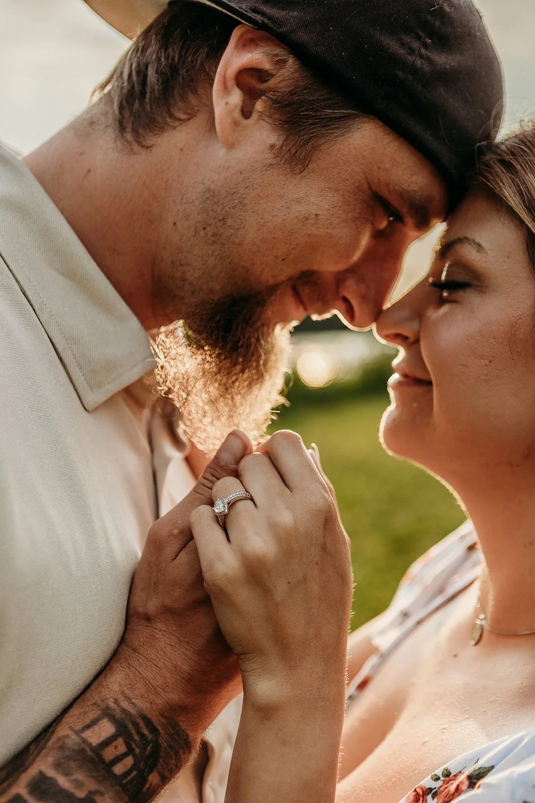 A couple touching foreheads and smiling with their eyes closed, holding hands with wedding rings, outdoors in warm sunlight.