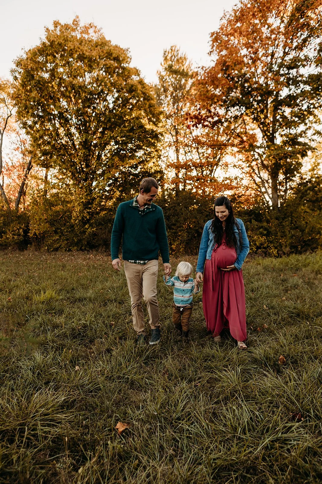 Family of three walking in a grassy field during autumn, with trees showing fall foliage in the background and warm sunset lighting.