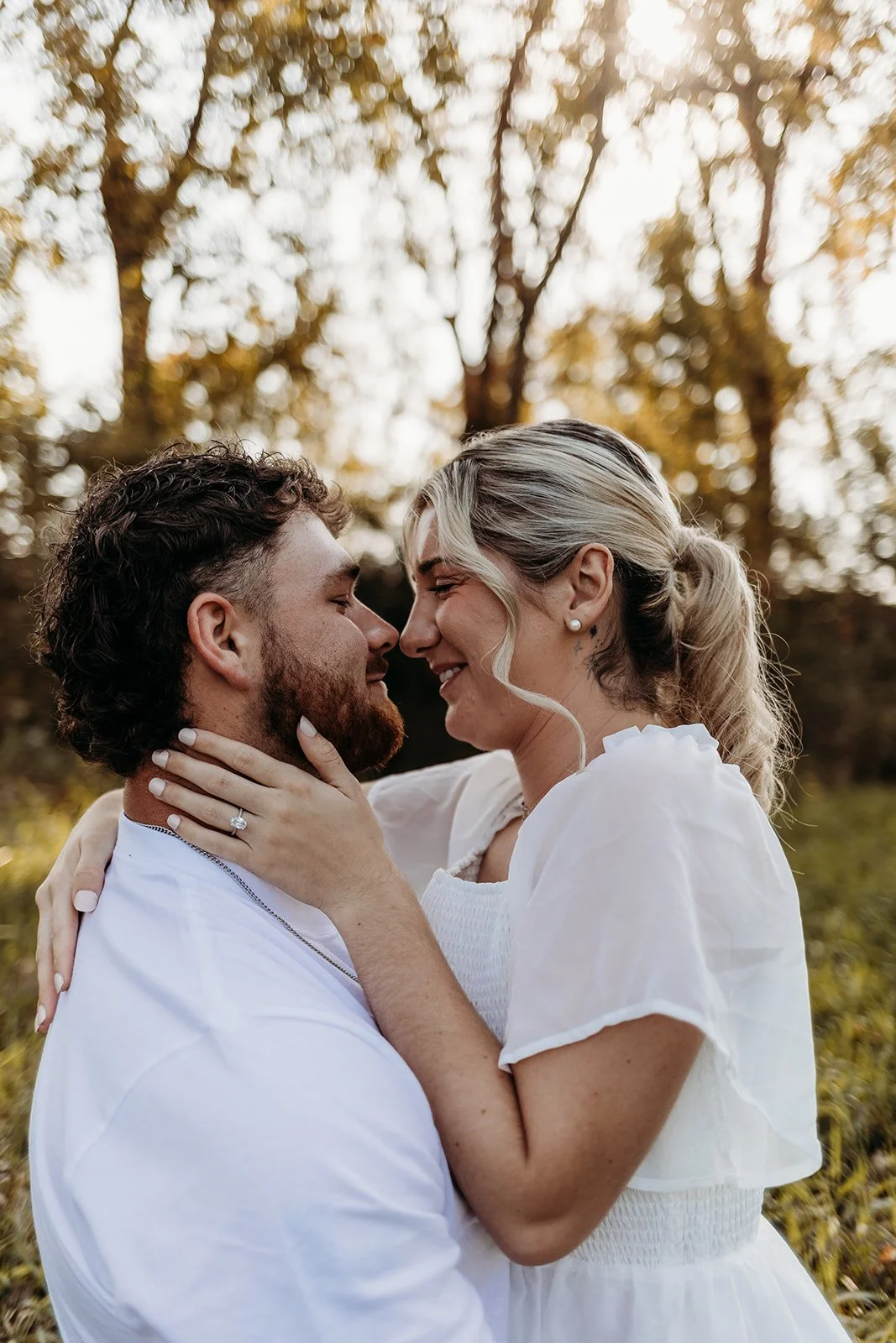 A couple in love face each other closely outdoors during autumn, with trees and fall foliage in the background.