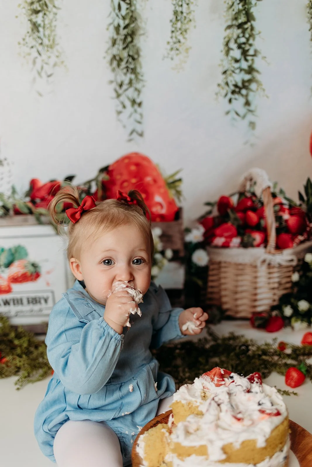 A young child with red bows in their hair, wearing a light blue shirt, eating cake with strawberry topping, with strawberries and strawberries in a basket in the background.