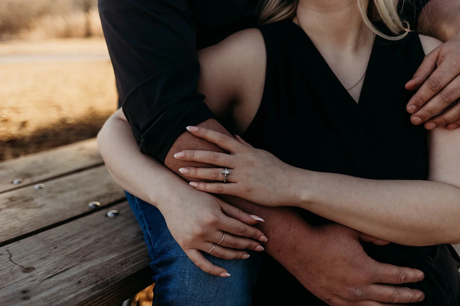 Close-up of a couple sitting on a wooden bench, with the woman wearing a diamond ring on her finger, and both embracing each other outdoors.