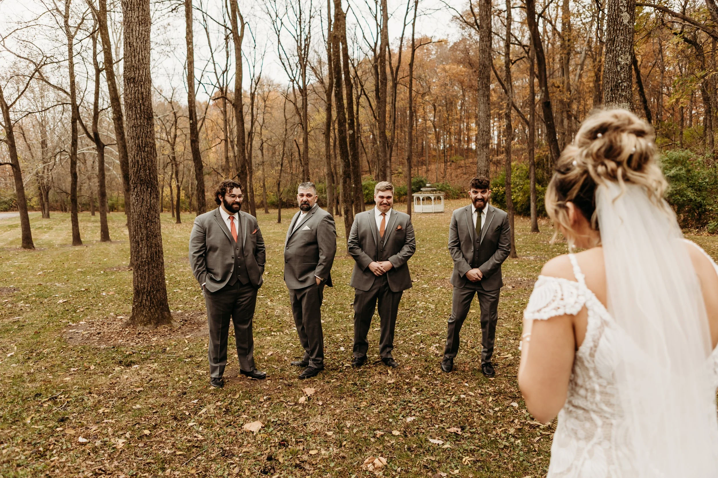 A bride facing four groomsmen outdoors during fall, with trees and a small white gazebo in the background.