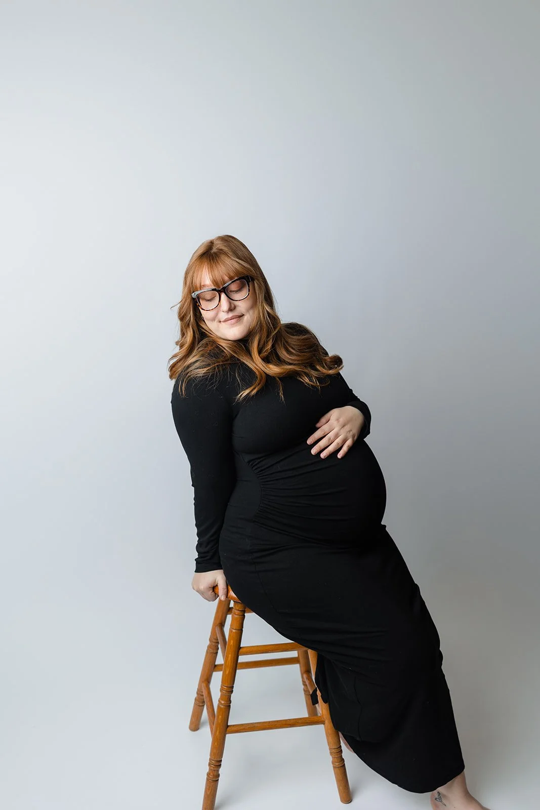 A pregnant woman with red hair and glasses, wearing a black dress, sitting on a wooden stool against a plain light gray background, with her eyes closed and one hand on her belly.