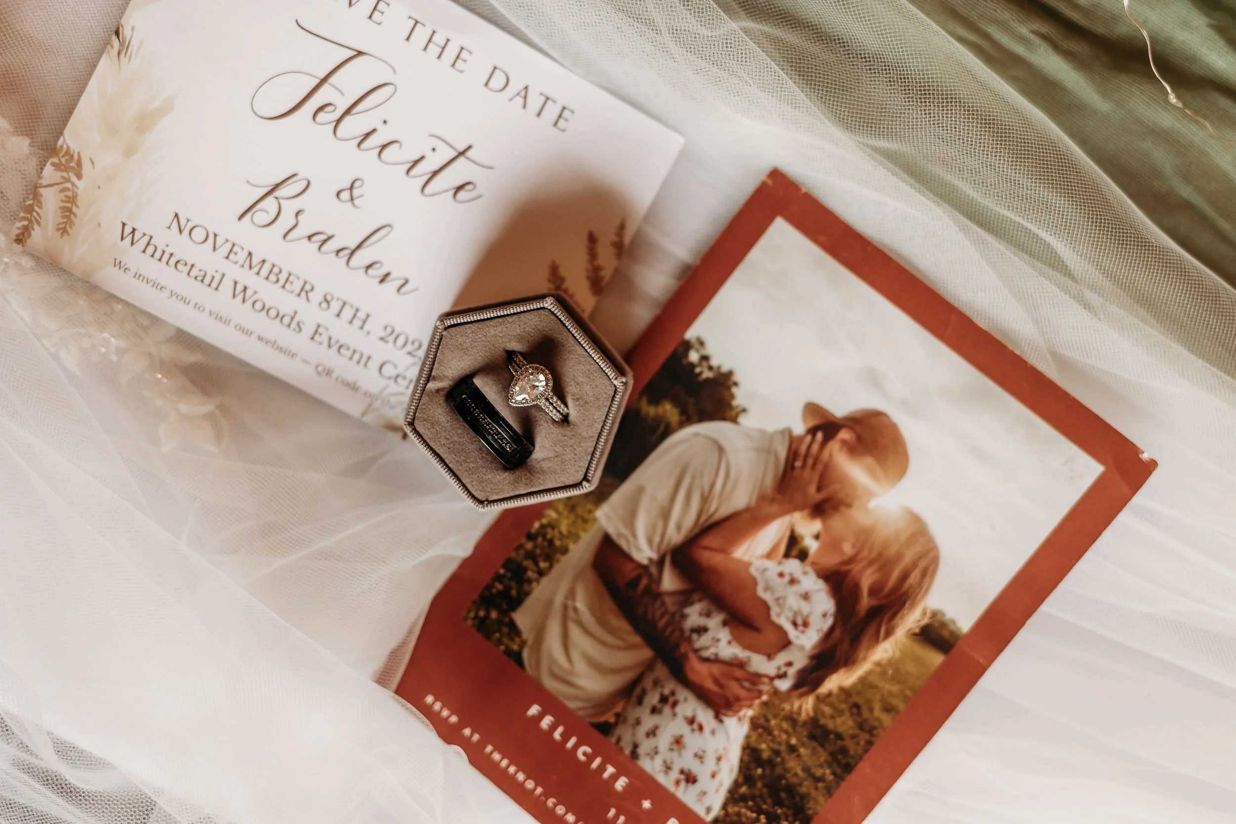Wedding invitation, a diamond engagement ring, a black wedding band, and a couple kissing in a photo, all laid out on white fabric.