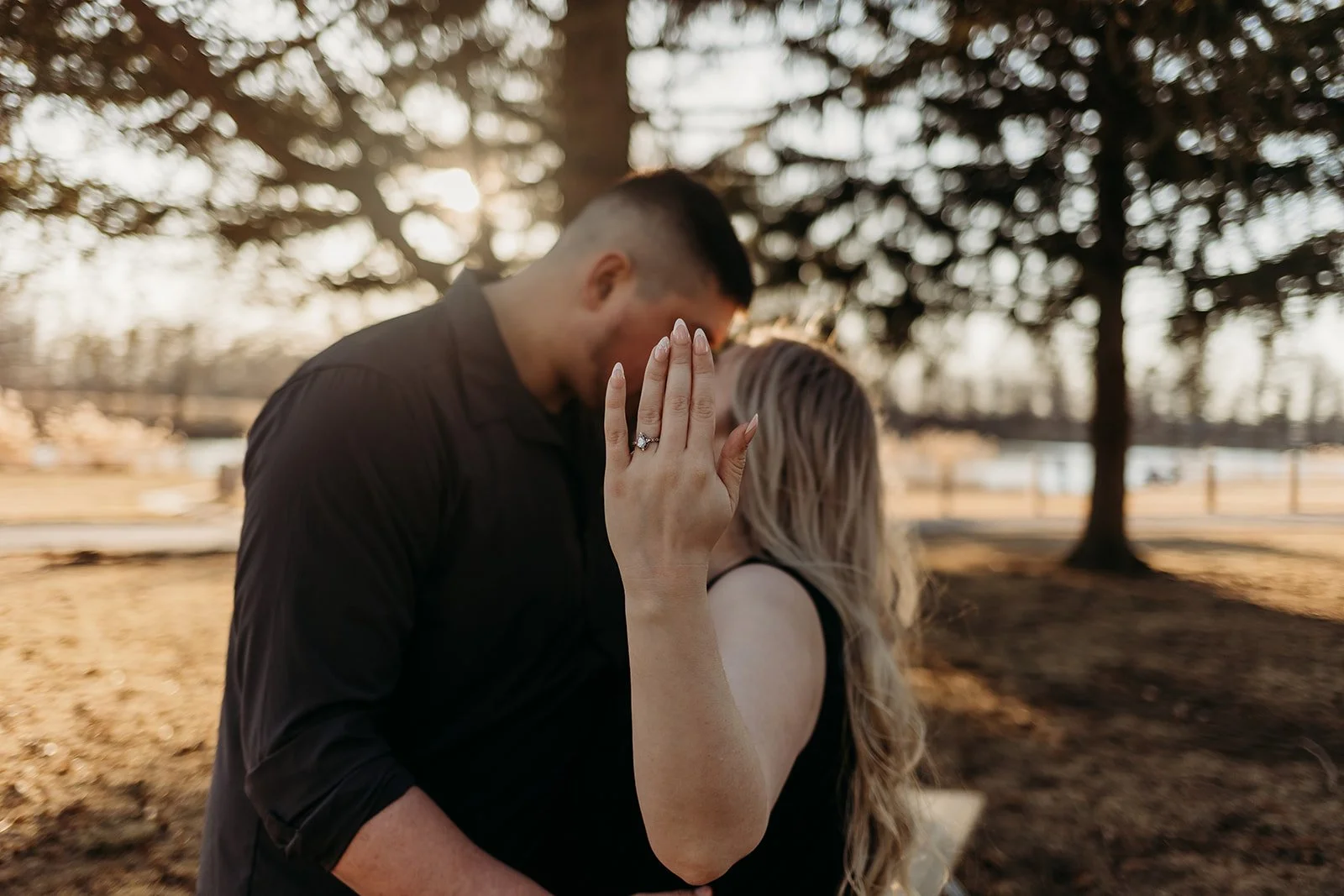 A couple in love sharing a kiss outdoors during sunset, with the woman showing her engagement ring.
