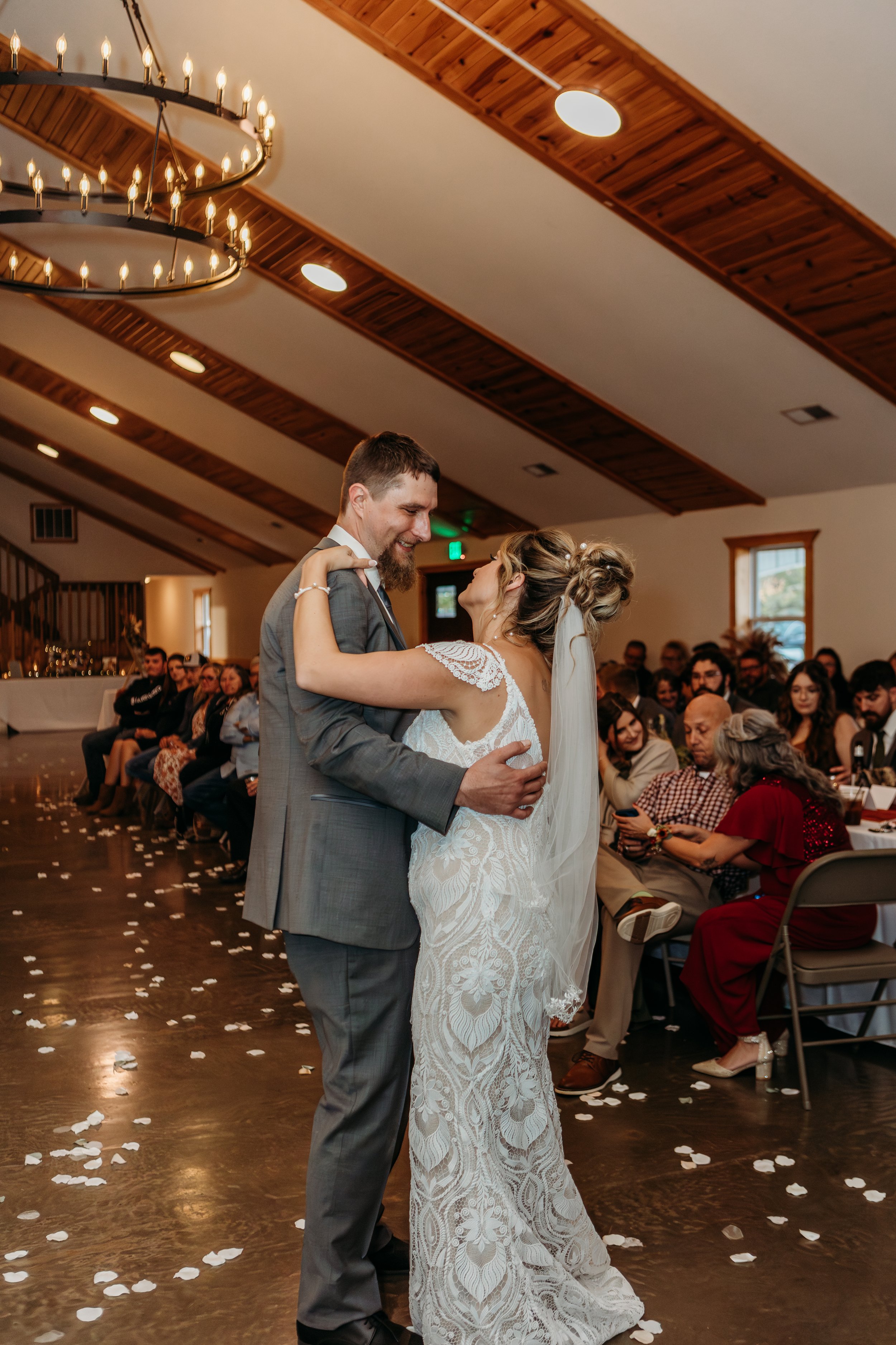 Bride and groom dancing at their wedding reception inside a banquet hall with friends and family seated at tables, wood-paneled ceiling, and a chandelier.