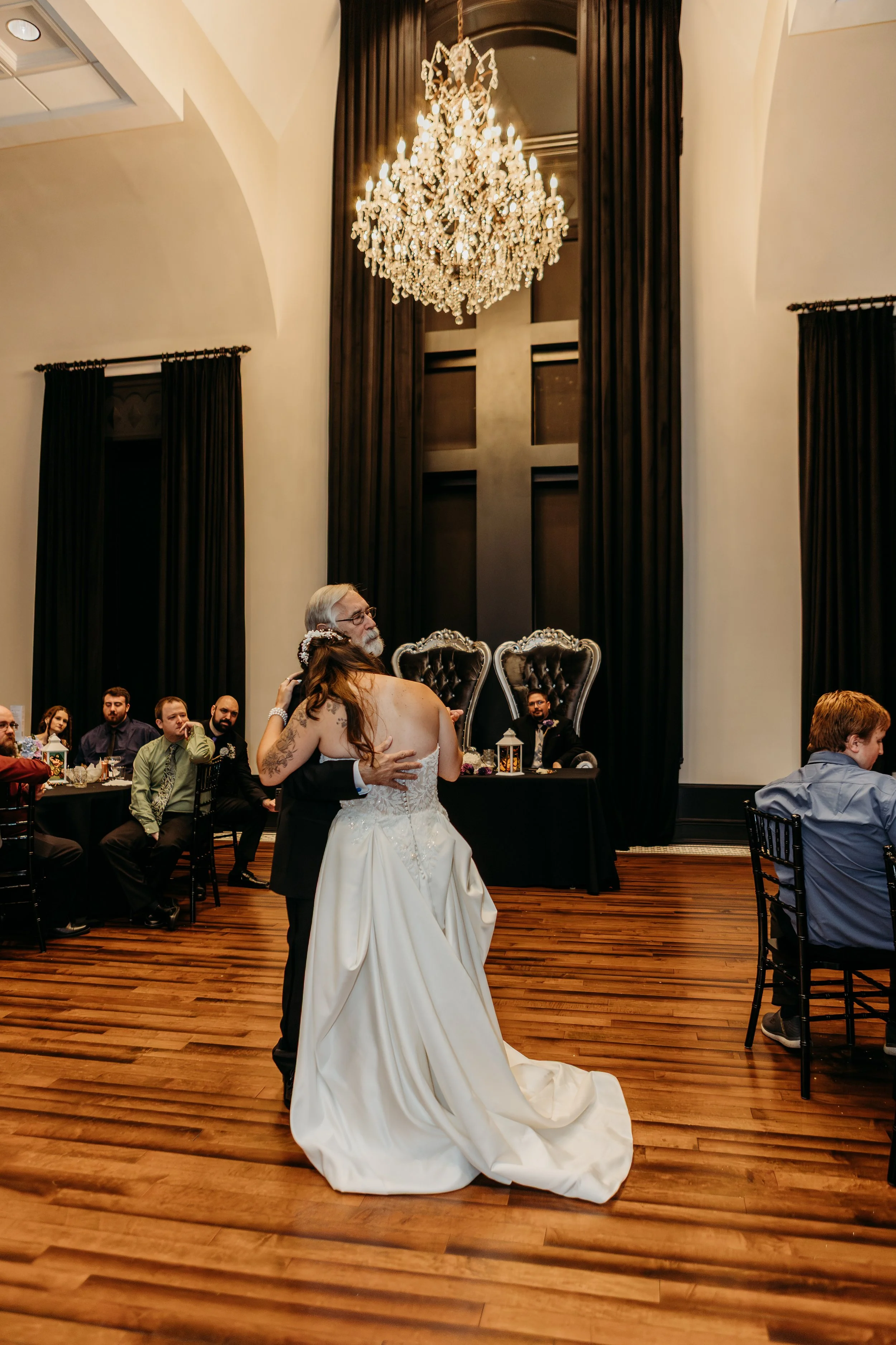 A wedding reception with a bride and groom sharing a dance in a grand hall with a high ceiling, chandelier, dark curtains, and guests seated at tables watching.