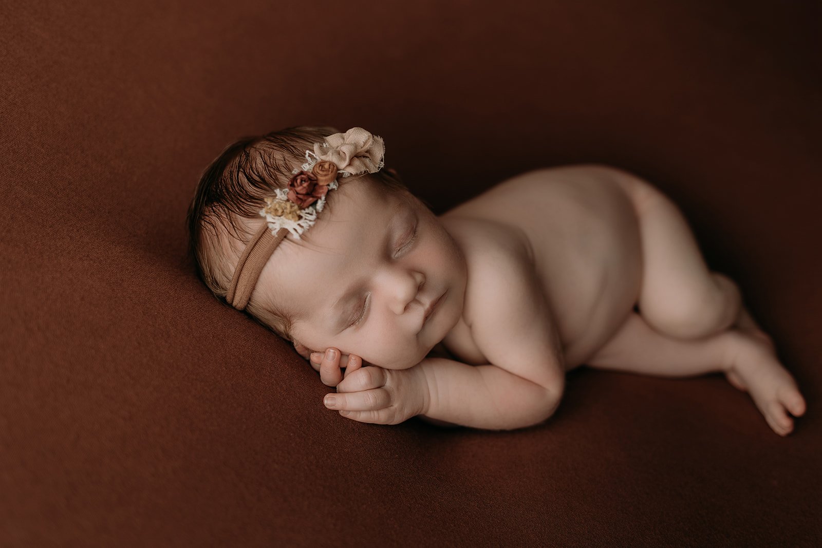 A sleeping newborn baby lying on a brown surface, wearing a beige headband with small flowers, and resting her face on her hand.
