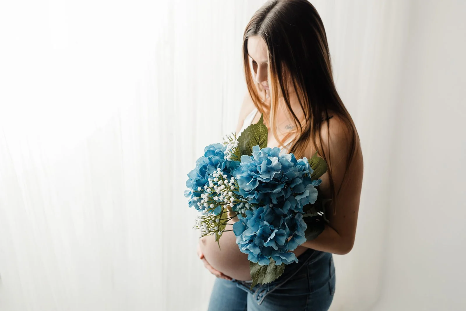 Pregnant woman holding a bouquet of blue hydrangeas and white flowers, standing against a white background.