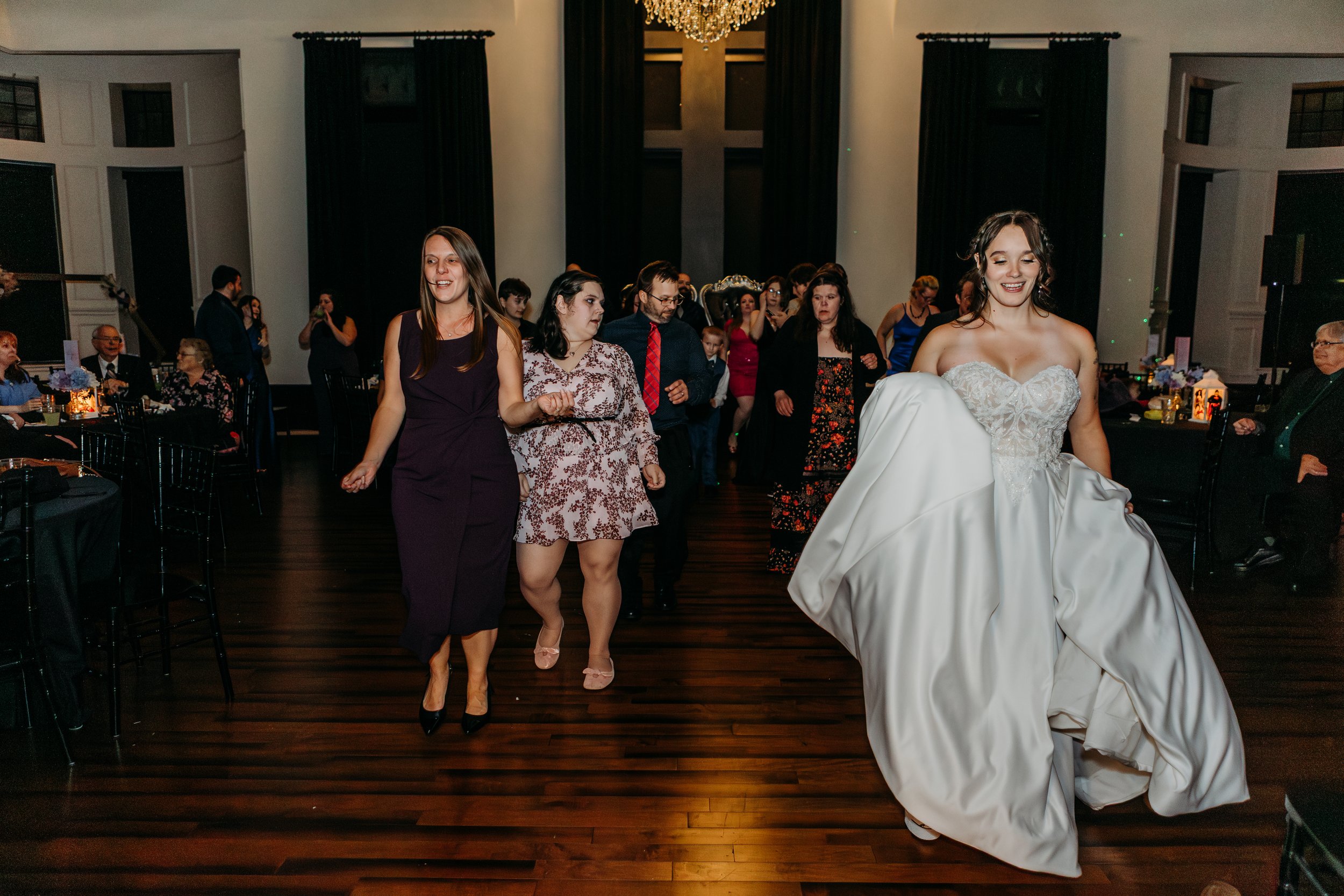 Bride in a strapless wedding gown dancing in a line with guests on a dark wooden dance floor at a wedding reception.