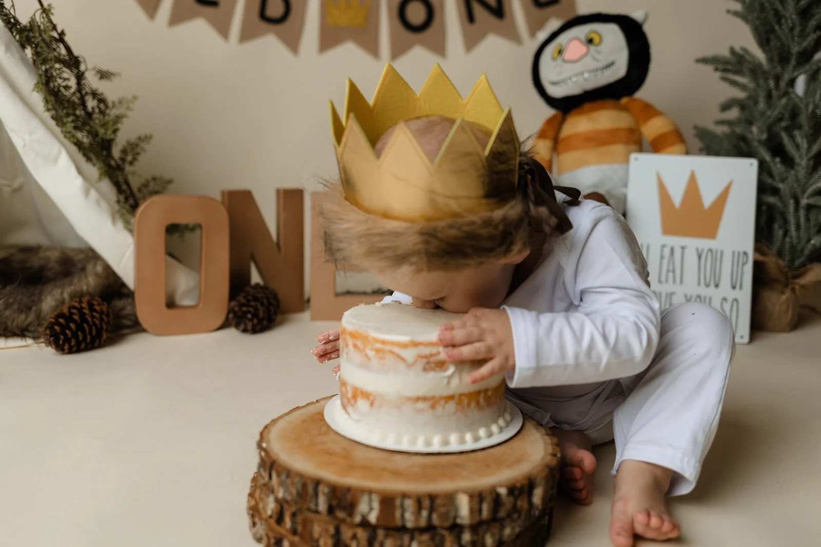 Child wearing a gold paper crown and a brown wig, leaning over a small, white frosted cake on a wooden slice, likely celebrating a birthday.