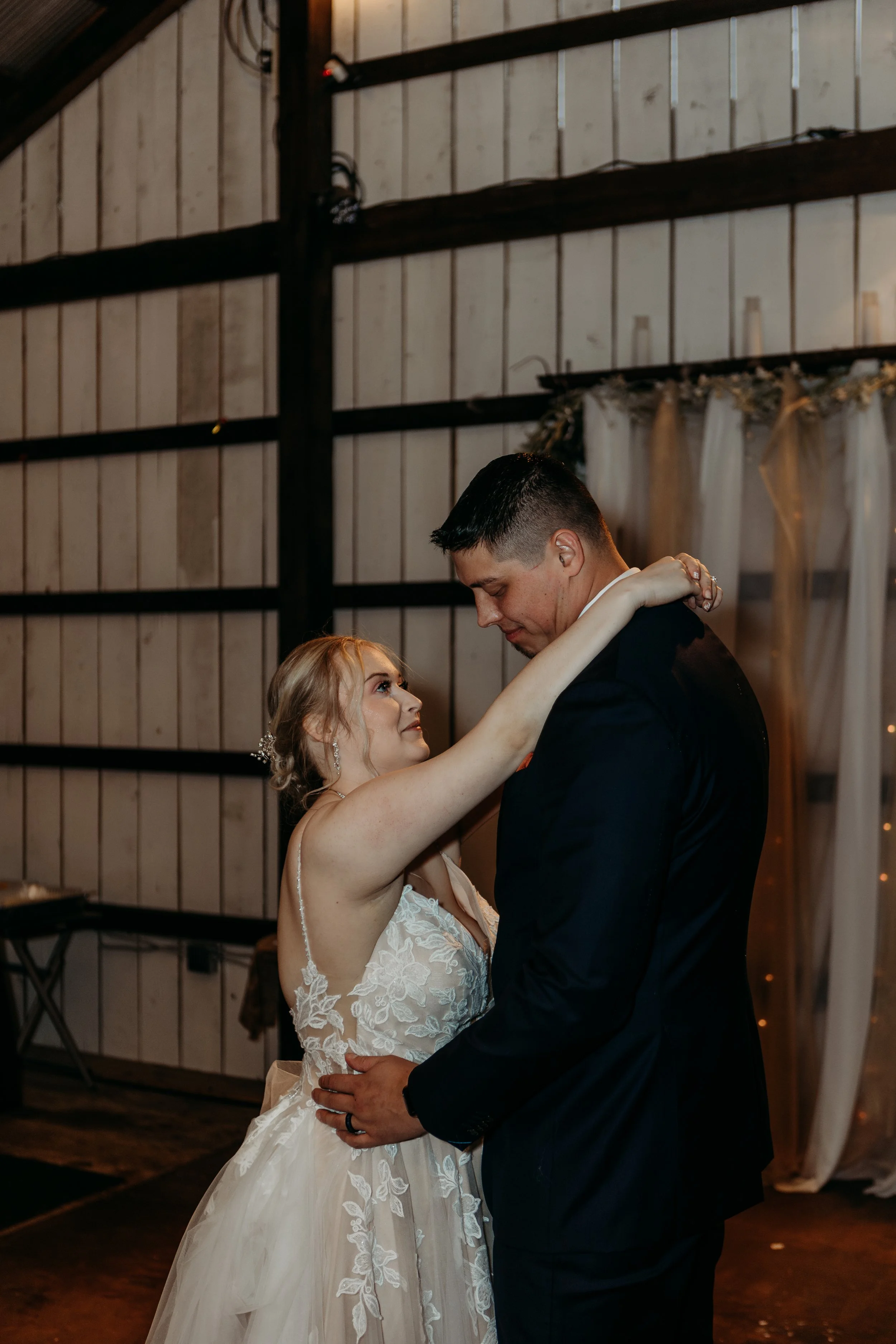 A bride and groom are dancing together at their wedding reception inside a barn. The bride is wearing a white lace wedding dress and has her arms around the groom's neck. The groom is wearing a dark suit and is holding the bride around her waist.