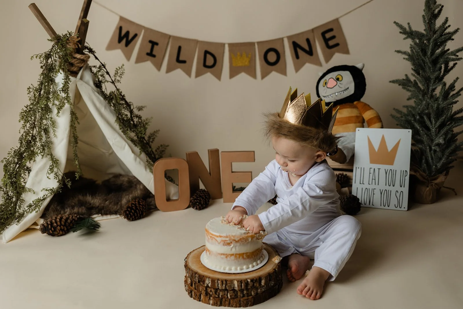 Child with a golden crown sitting on the floor, smashing a small white cake on a wooden slab, celebrating a first birthday with a wilderness-themed setup, including a teepee, pinecones, a stuffed animal, and a "WILD ONE" banner.