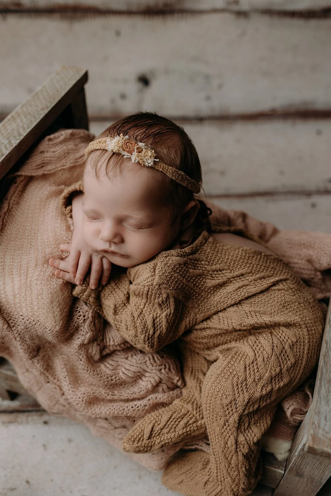 A sleeping baby wrapped in brown knitted blankets with a floral headband, lying on a soft surface near a rustic wooden wall.