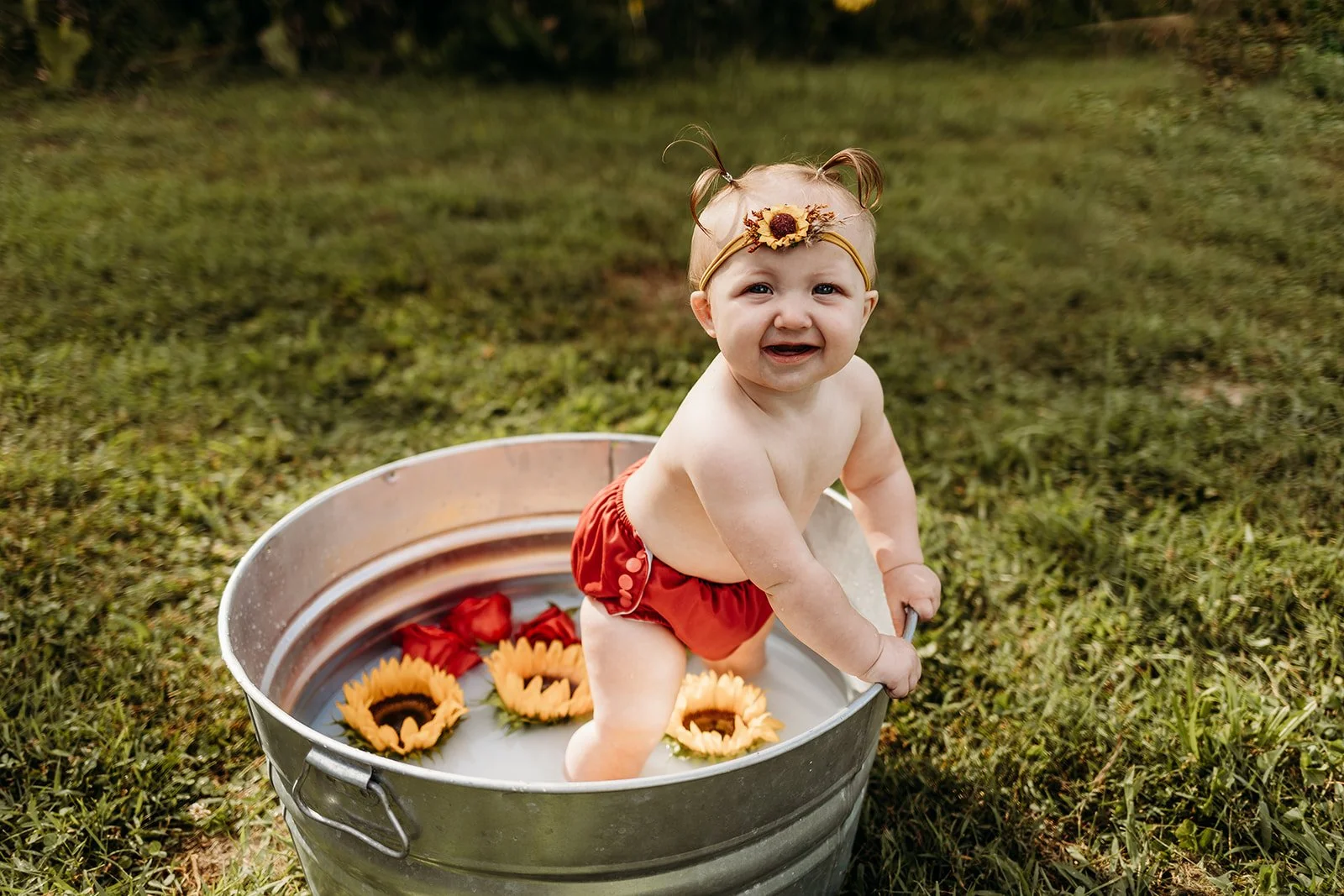 A baby with red shorts, a sunflower headband, and no shirt, sitting in a metal tub filled with water and floating flowers, outdoors on grass.
