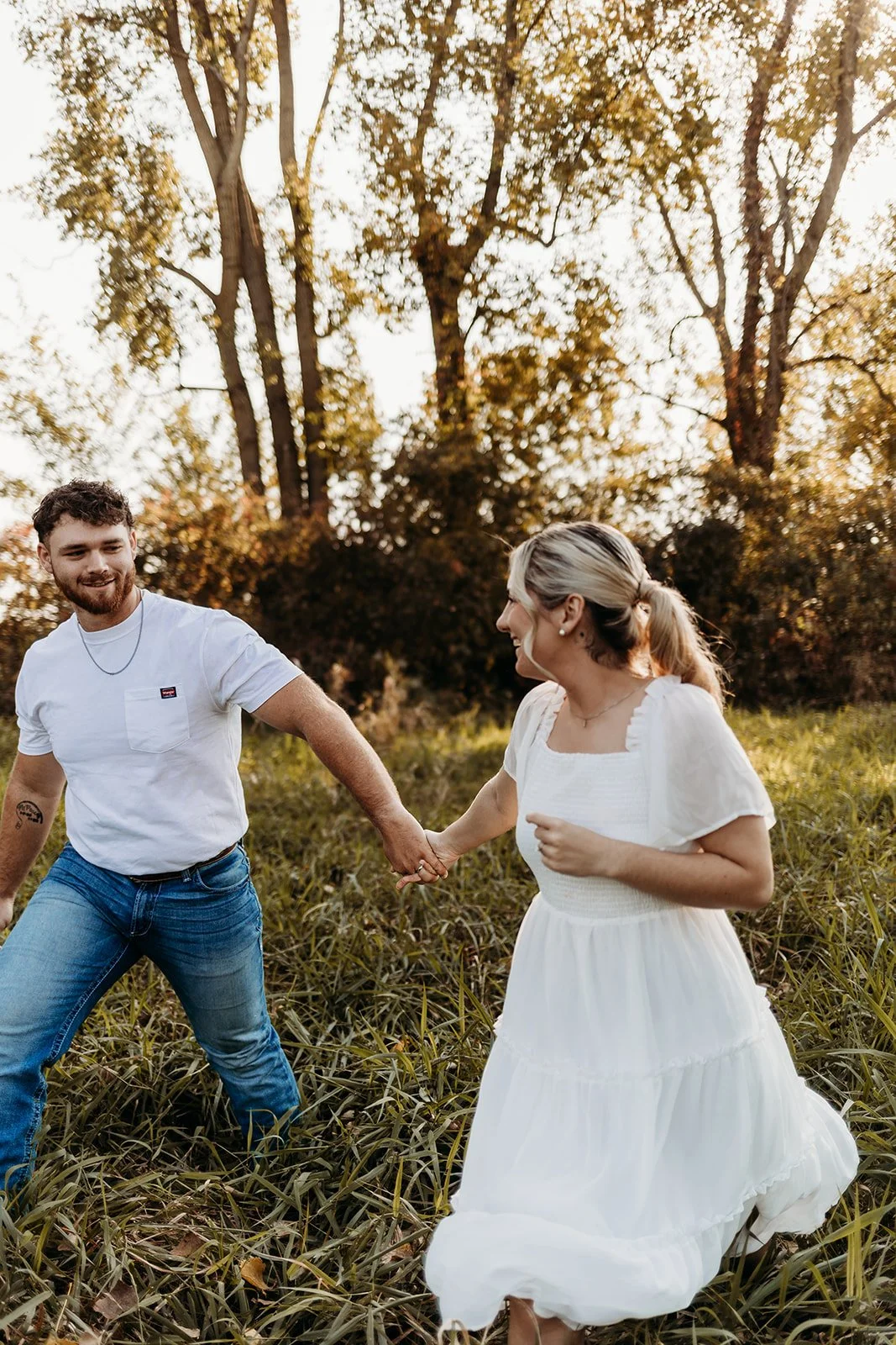 A couple holding hands and dancing outdoors in a grassy field with trees and warm sunlight in the background.