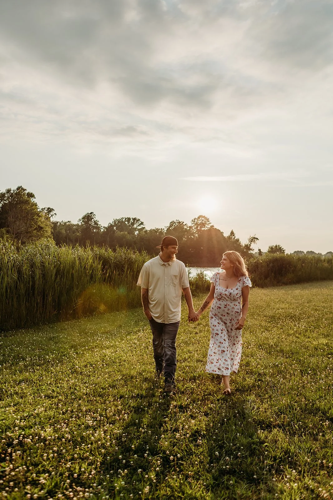 A couple walking hand in hand through a grassy field near a lake at sunset, with trees in the background and a cloudy sky overhead.