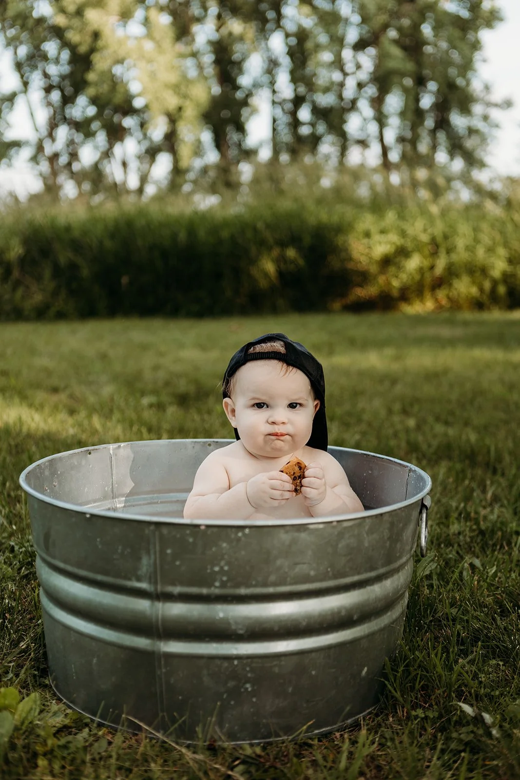 A baby sitting in a metal tub filled with water outdoors, wearing a backwards cap and holding a cookie.