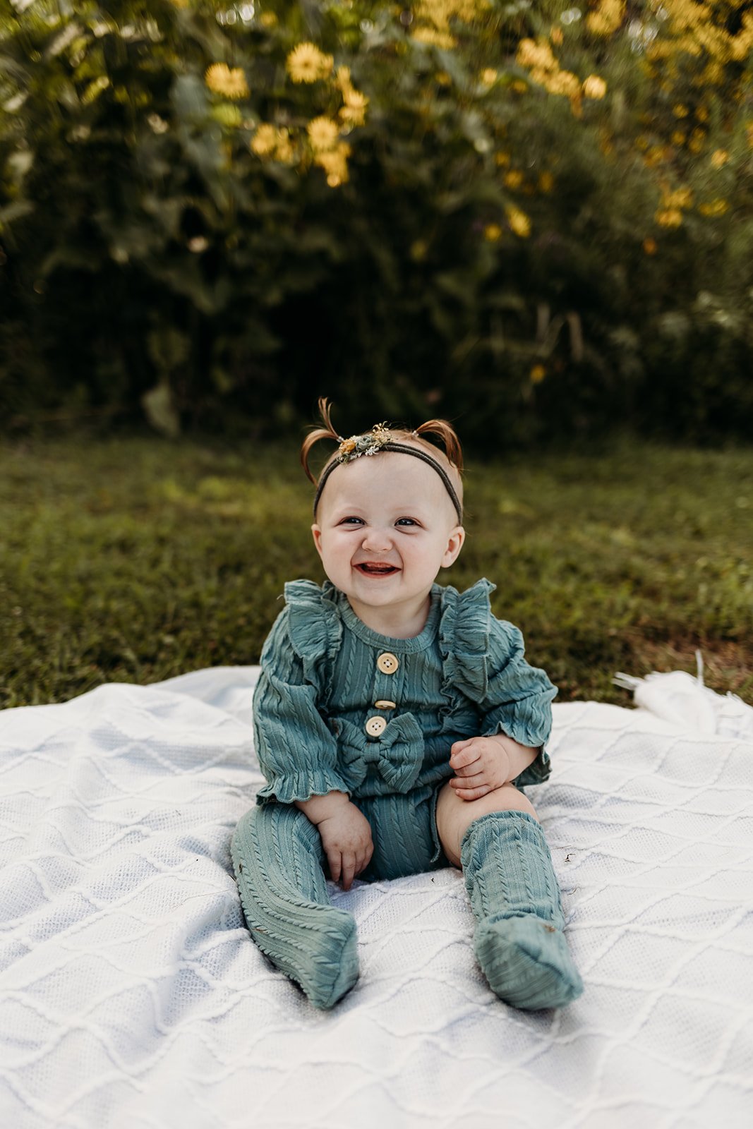A smiling baby girl sitting on a white blanket outdoors with greenery and yellow flowers in the background.