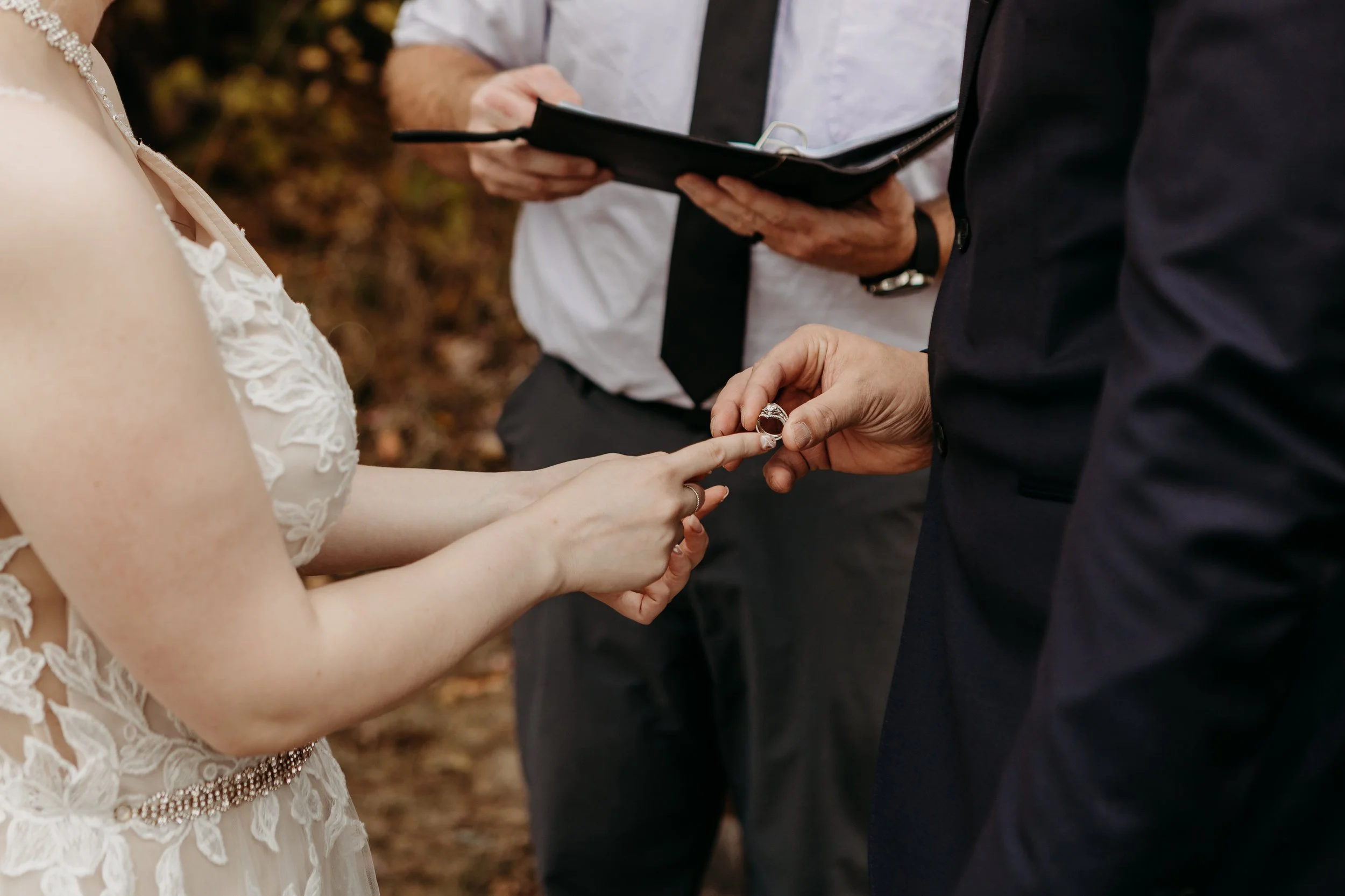 A bride and groom exchanging wedding rings outdoors, with an officiant holding a book in the background.
