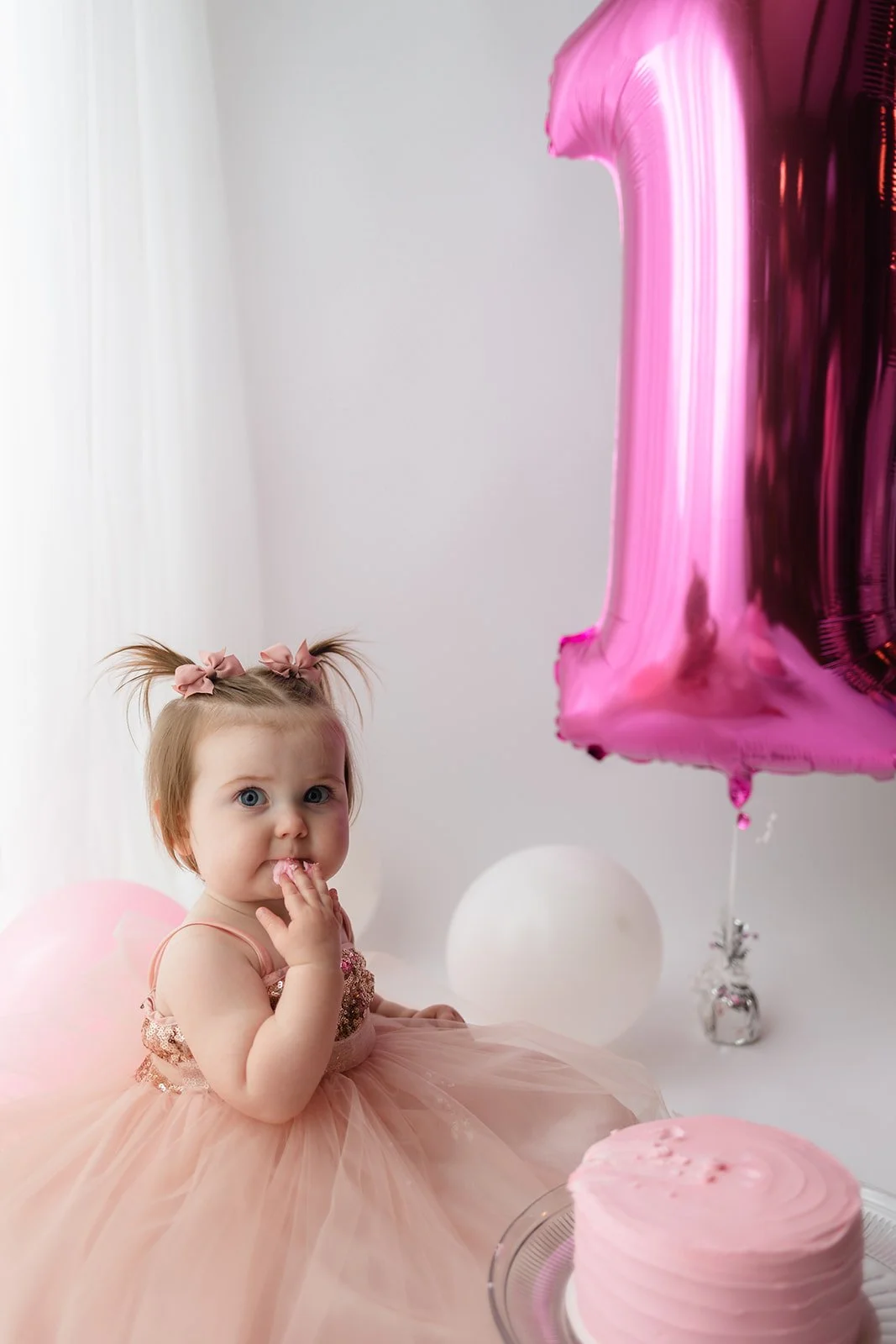 A young girl in a pink dress with puffy hair tied with pink bows sitting at her birthday celebration with pink and white balloons, a pink cake, and a large pink number one balloon.
