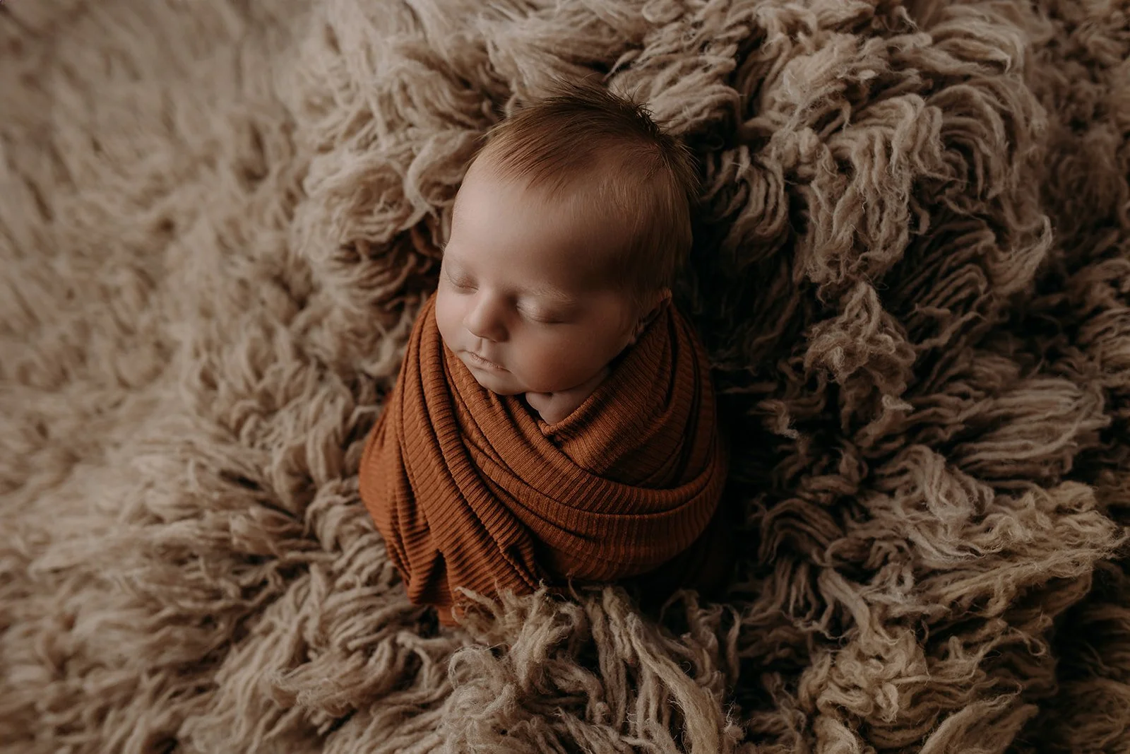 A sleeping baby wrapped in a brown blanket, lying on a soft, furry beige rug.