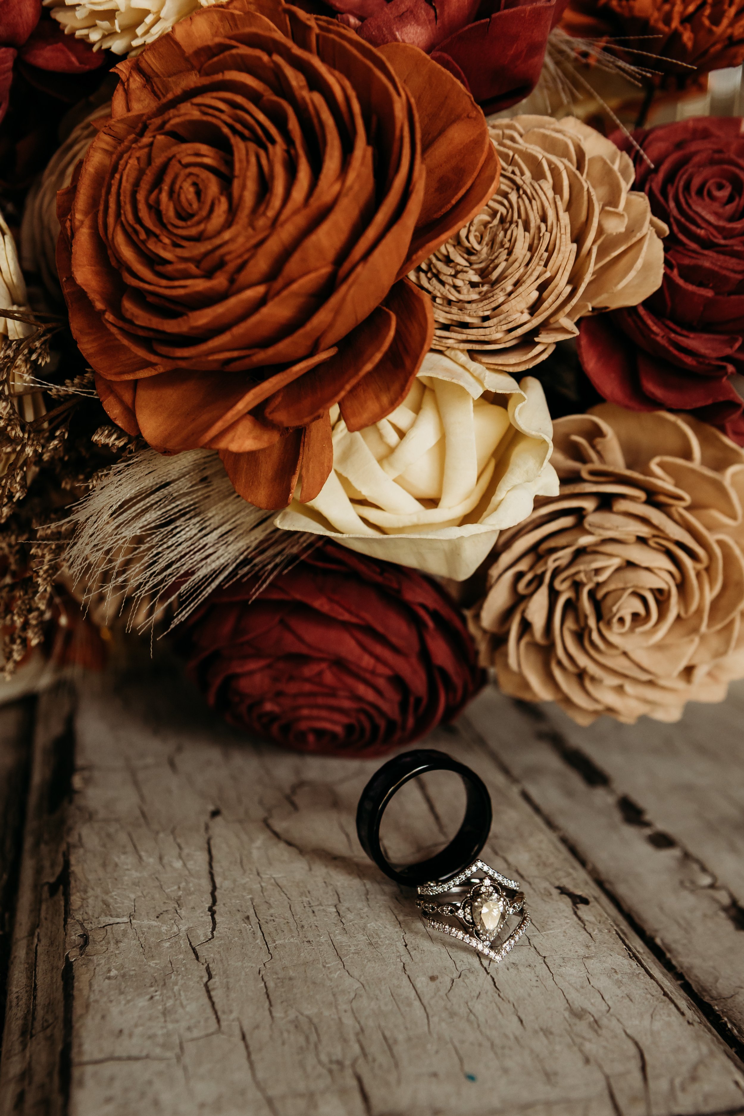 A bouquet of artificial roses in shades of brown, cream, and red, with a feather and dried flowers, placed on a rustic wooden surface. Two rings, one black band and one with a large gemstone, are positioned near the flowers.