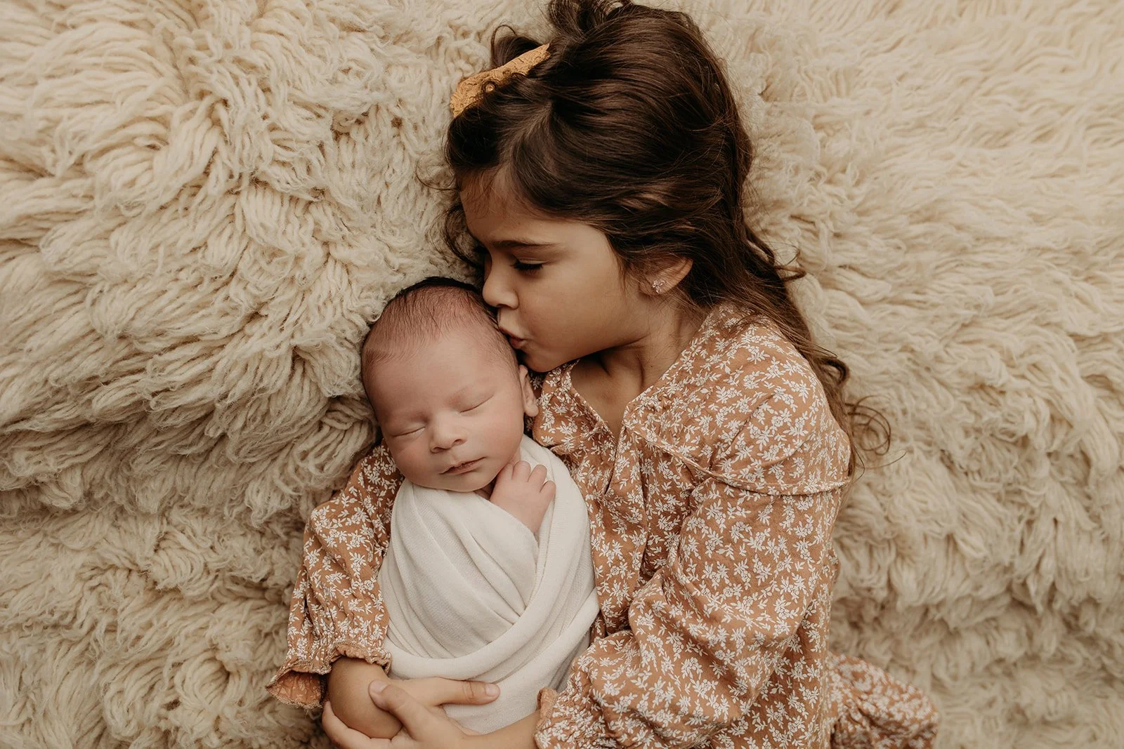 A young girl with long brown hair and a beige flower hairclip gently kisses a newborn baby on the forehead while lying on a fluffy cream-colored blanket.